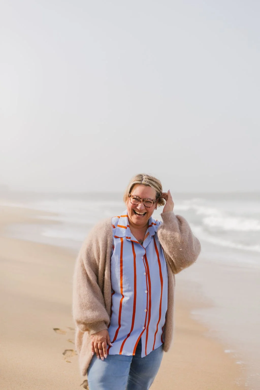 A woman with glasses and blonde hair smiling on a beach, wearing a striped shirt, beige cardigan, and blue jeans.