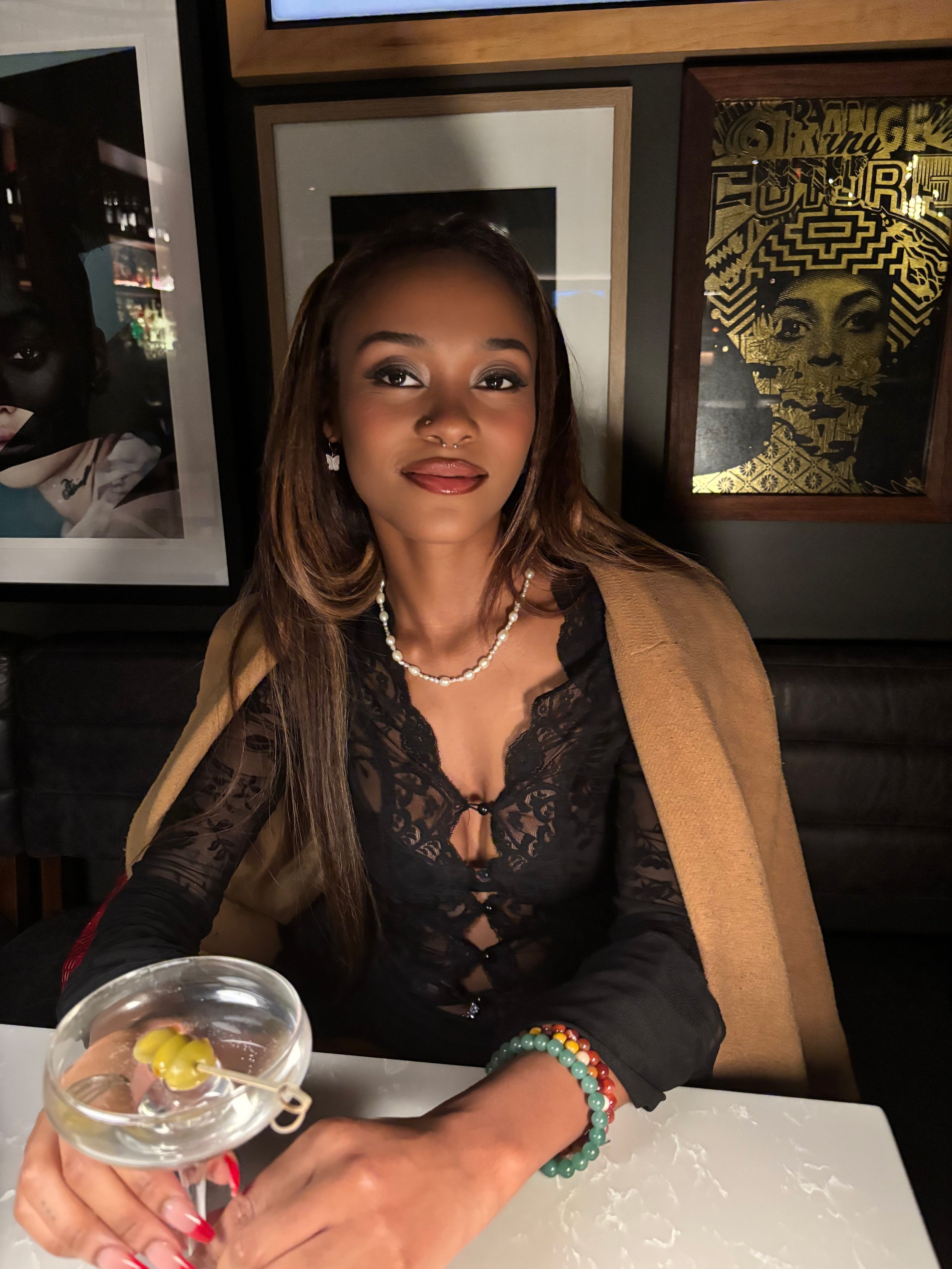 A young woman with light brown hair, wearing a black lace top, a pearl necklace, and colorful beaded bracelet, holding a martini glass with green olives, sitting at a table in a restaurant or bar with framed art on the wall behind her.