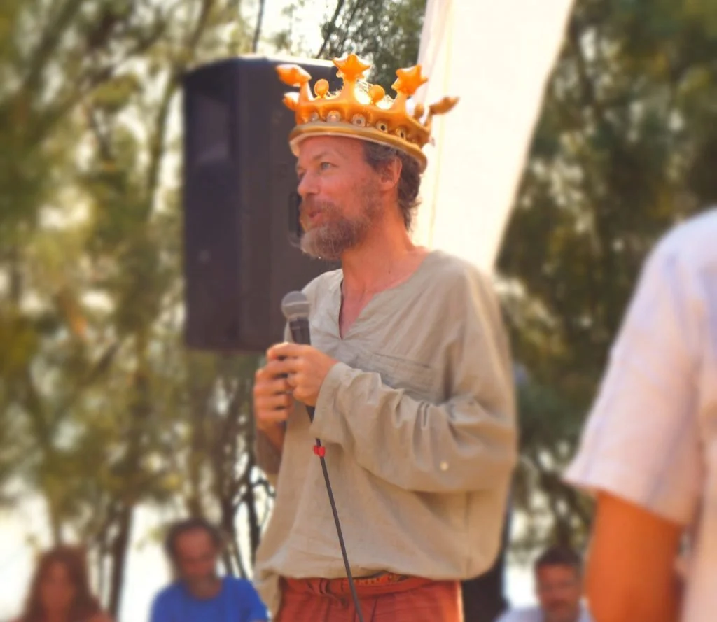 A man wearing a crown and gray shirt holding a microphone at an outdoor event organised by Poveglia per tutti on the island of Poveglia, in the Venice lagoon