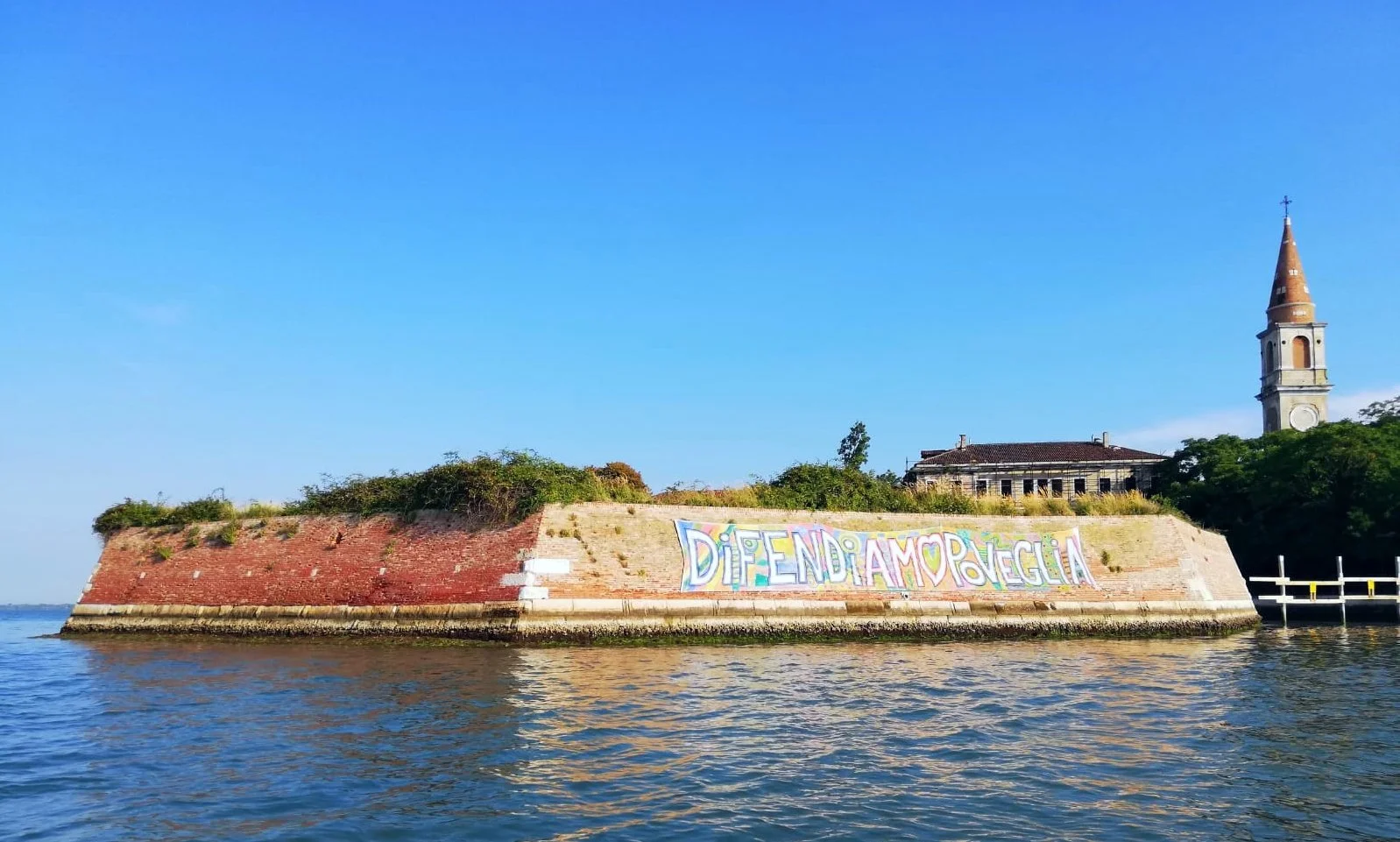 An image of the island of Poveglia in the Venice lagoon with a colorful graffiti message on its wall, and a church steeple visible in the background.