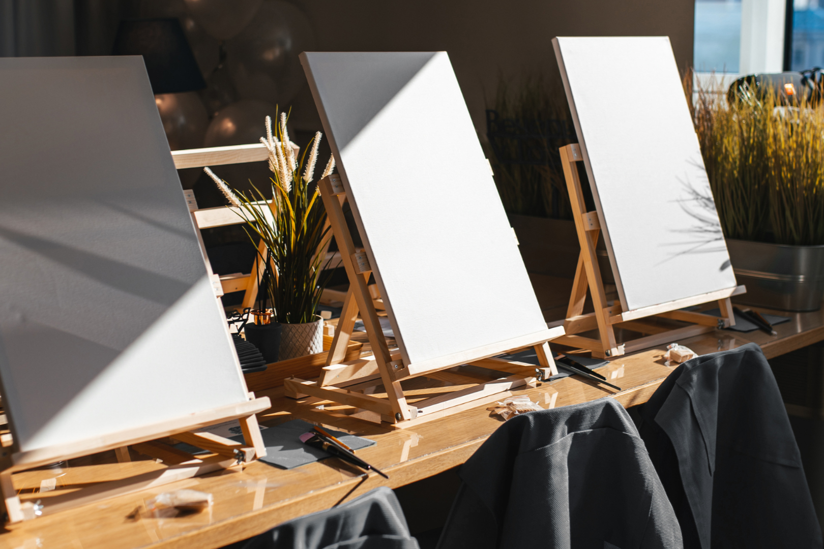 Empty canvas easels set up on a wooden table in a well-lit room, with paint brushes and plant decorations.
