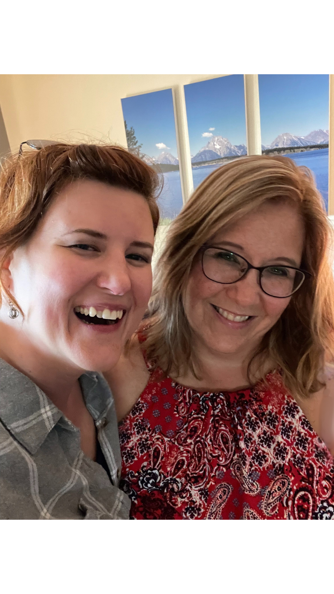 Two women smiling and taking a selfie indoors, with a mountain landscape visible through a triptych window behind them.