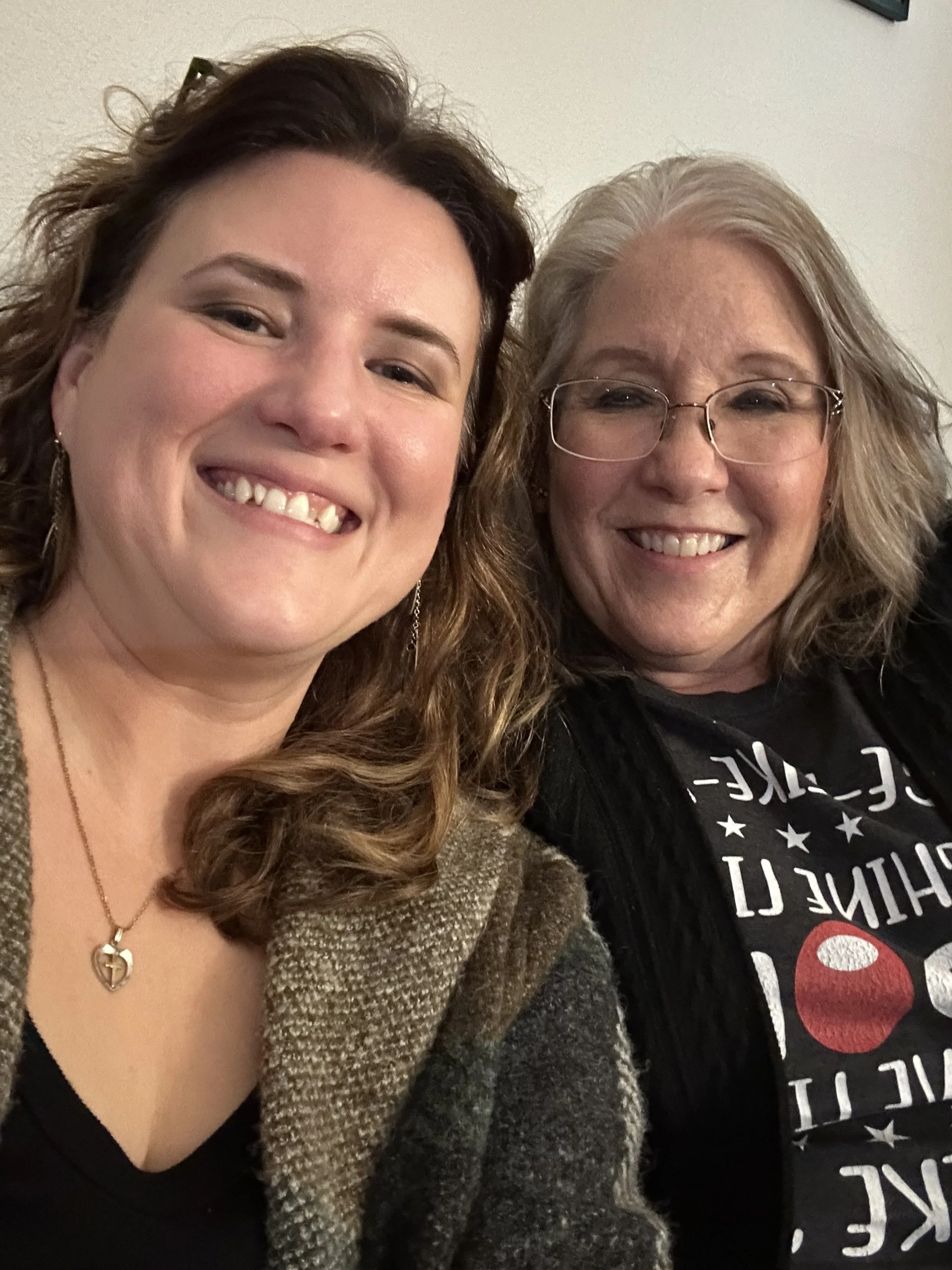 Two women smiling for a selfie, one with long curly brown hair, wearing a necklace with a heart-shaped pendant and a patterned coat, the other with shoulder-length gray hair, glasses, and a black shirt with white text and red graphic design.