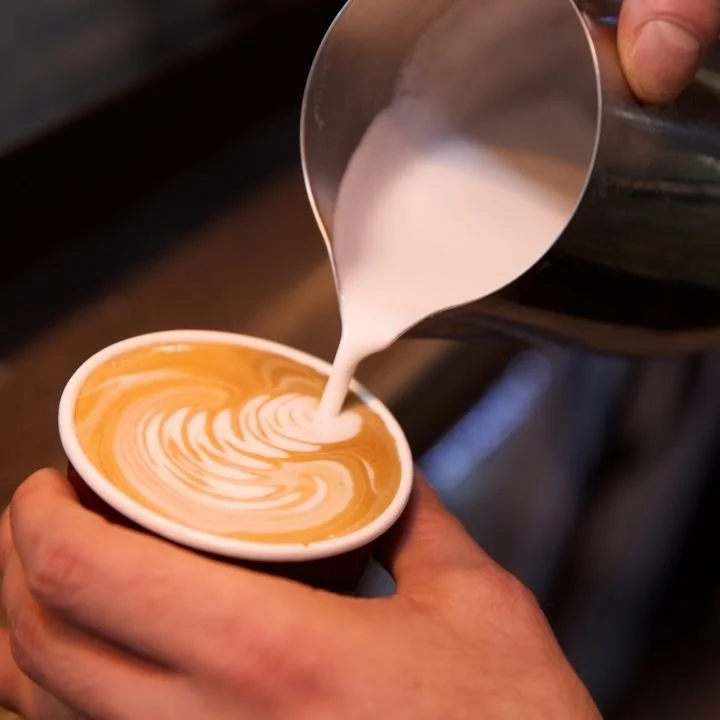 Barista pouring steamed milk into coffee, creating latte art pattern.