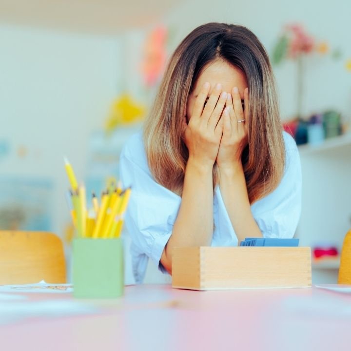 Stressed teacher covering her face with both hands at her desk.