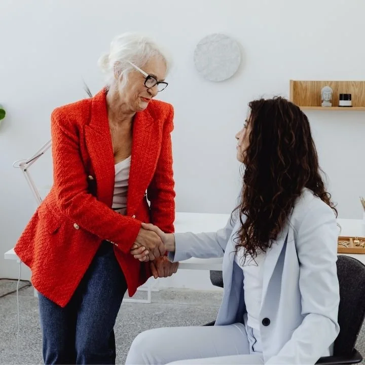 Older woman in red blazer shaking hands with younger professional in office.