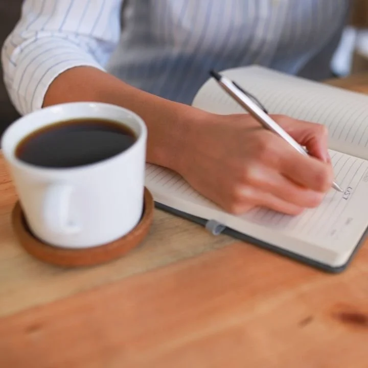 Hand writing in a notebook next to a cup of black coffee on a wooden table.