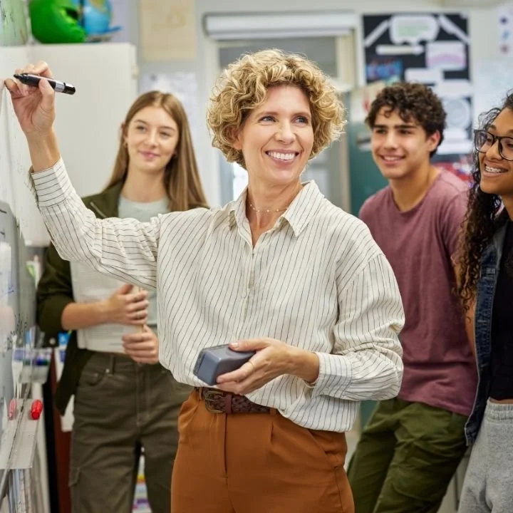 Teacher smiling while writing on classroom whiteboard with students gathered around.