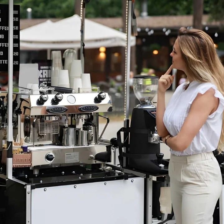 Person standing beside a mobile coffee station with espresso machine and grinder outdoors.