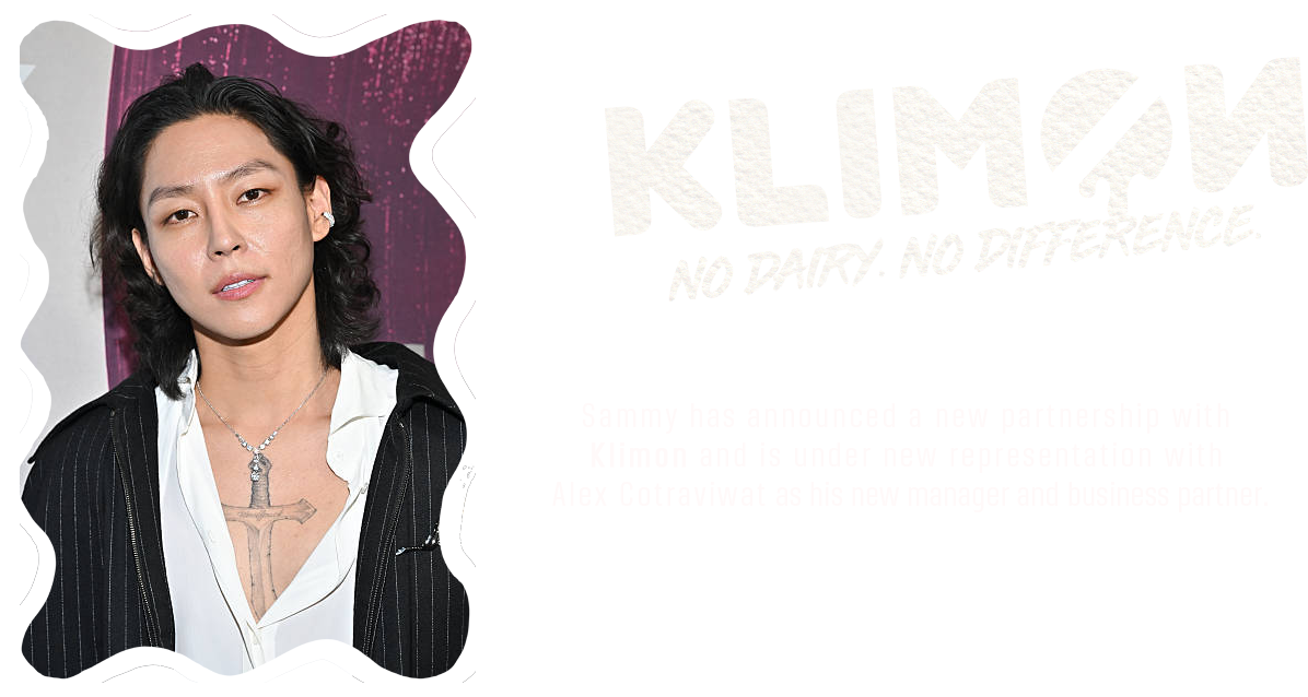 A person with shoulder-length black hair, wearing a black pinstripe blazer over a white shirt, and a prominent necklace with a sword pendant. The background features pink and purple abstract patterns.