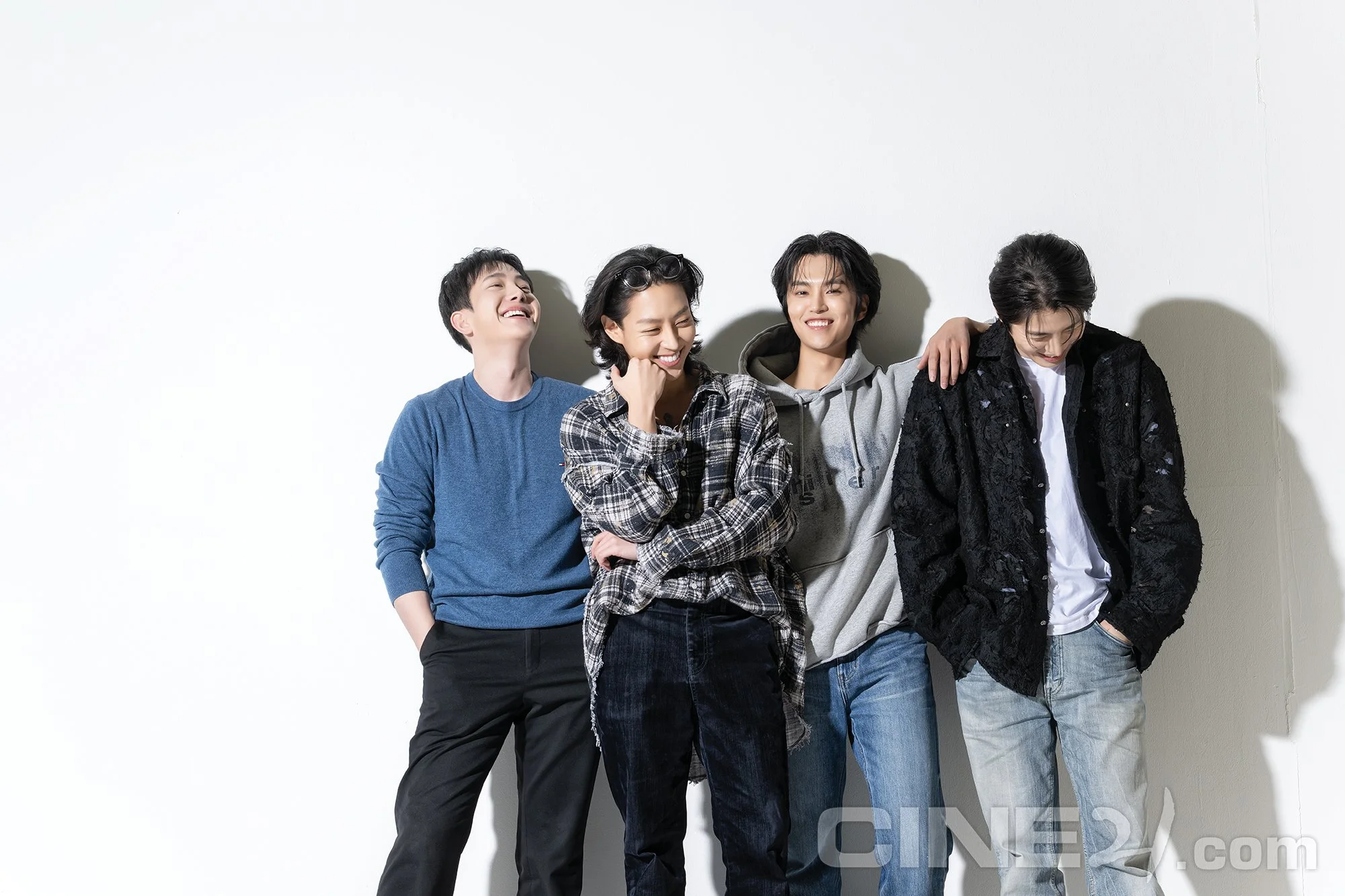 Four young men standing against a plain white wall, smiling and laughing, casually dressed in hoodies, t-shirts, and jeans.
