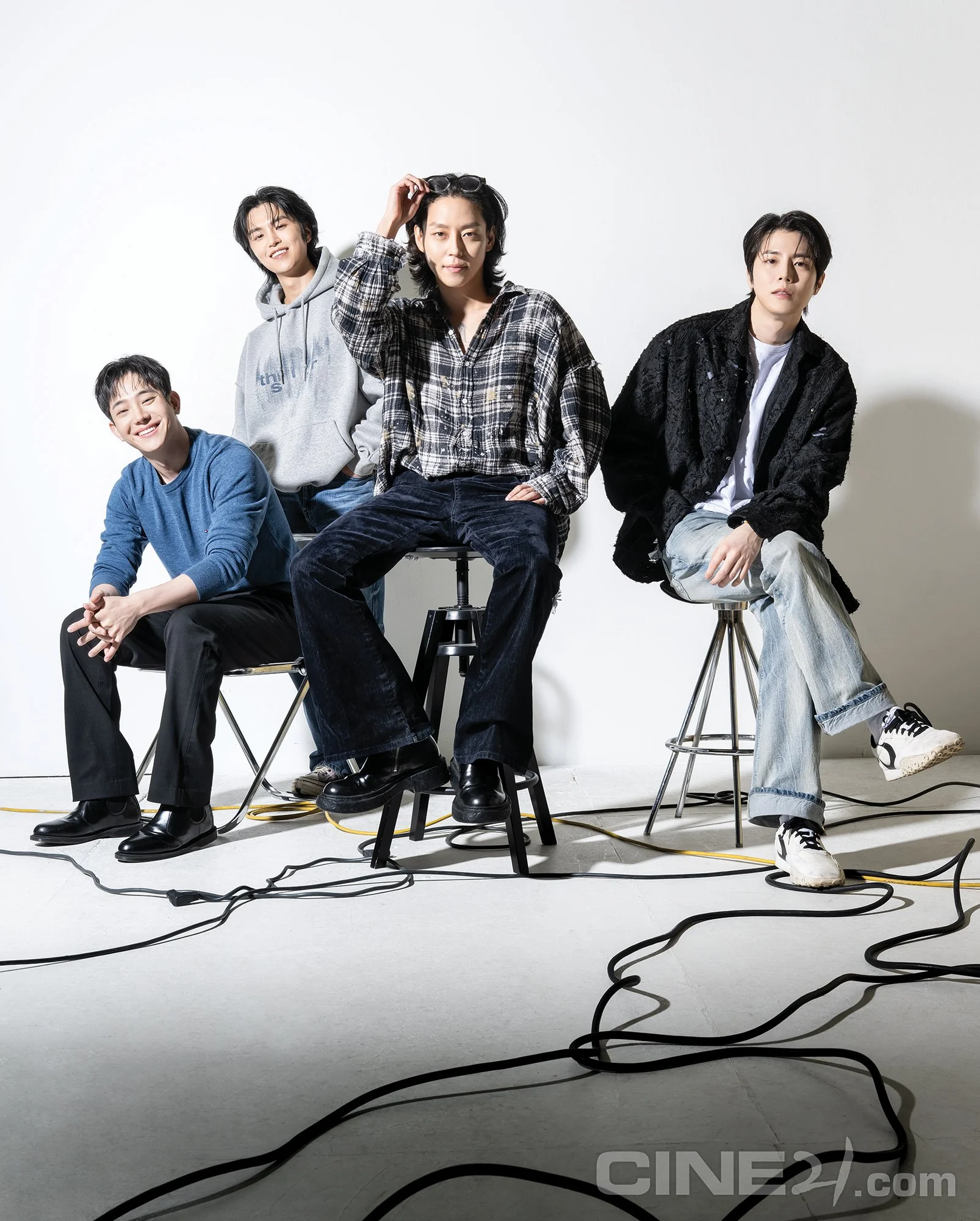 Four members of The Rose sitting and standing in a studio with a white background, surrounded by black electrical cords, dressed in casual clothing, posing for a photo.