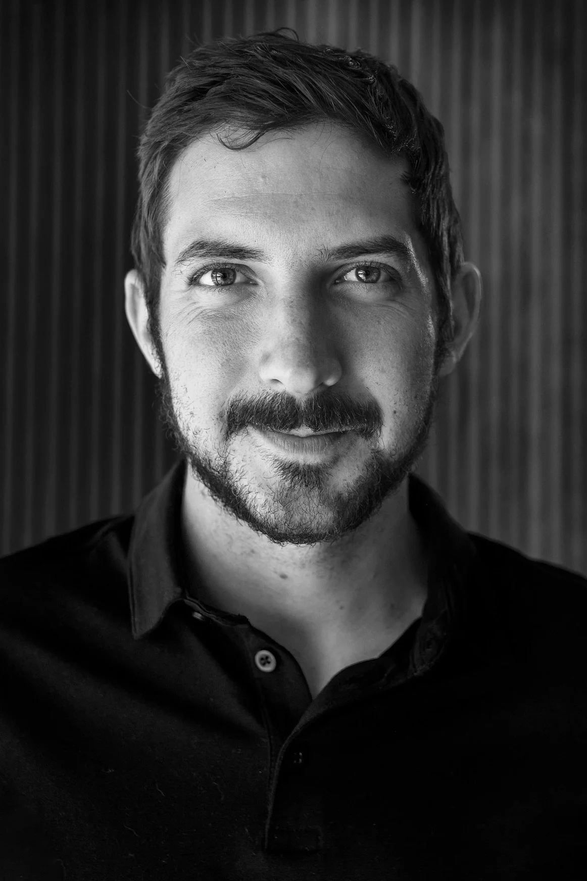 Black and white portrait of a man with short dark hair and a beard, wearing a dark polo shirt, smiling slightly, standing in front of a textured background.