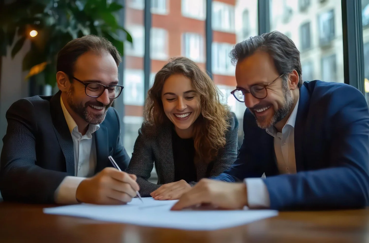 Tres personas de negocios sentados en una mesa, riendo y firmando un documento, en un ambiente de oficina con ventanas grandes.