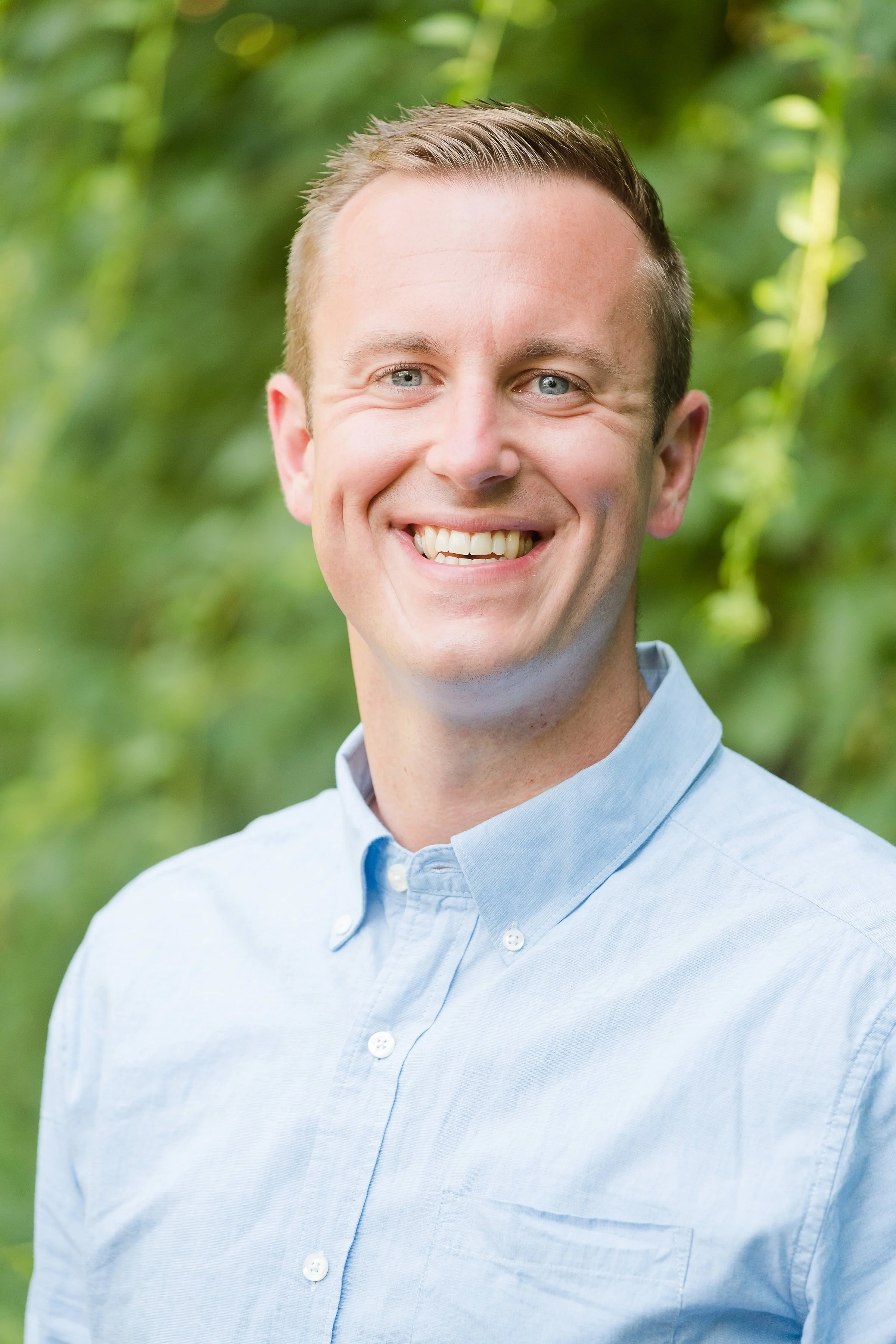 A smiling man in a light blue button-up shirt outdoors with green foliage in the background.