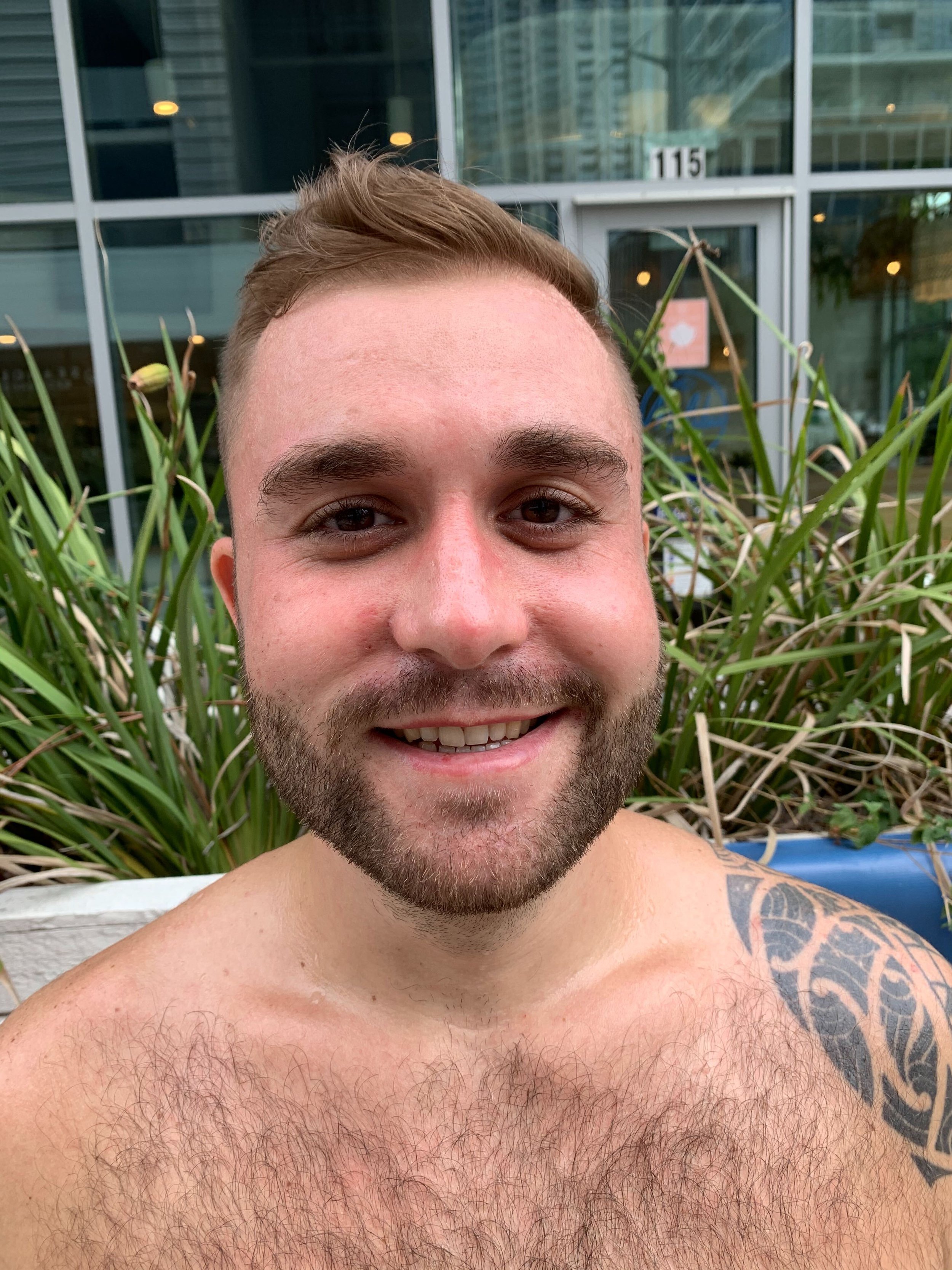 A smiling man with a beard and short hair taking a selfie outdoors, with plants and a modern glass building in the background.