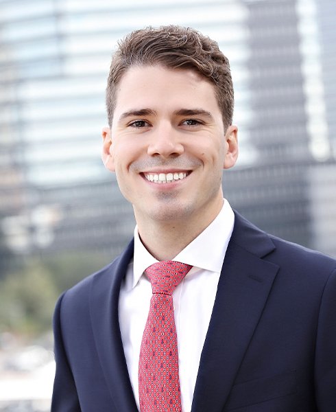 A young man with short brown hair, wearing a navy suit, white shirt, and pink patterned tie, smiling outdoors with a cityscape in the background.
