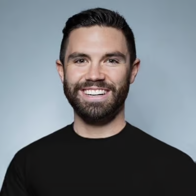 A smiling man with short dark hair, a beard, wearing a black shirt, against a light gray background.