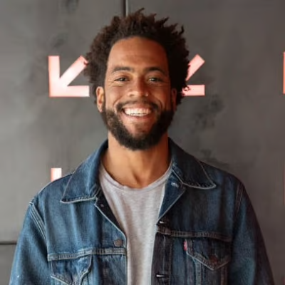 Smiling man with curly hair and beard wearing a denim jacket and gray T-shirt, standing in front of a dark background with pink geometric shapes.