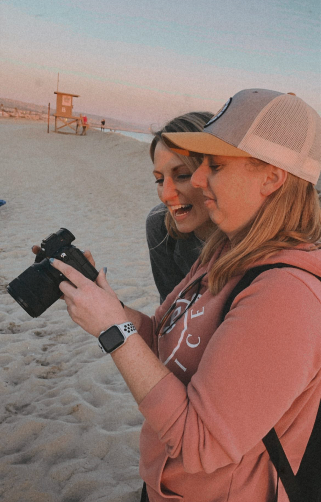 Two smiling women on the beach looking at a camera, with a lifeguard tower and sunset in the background.