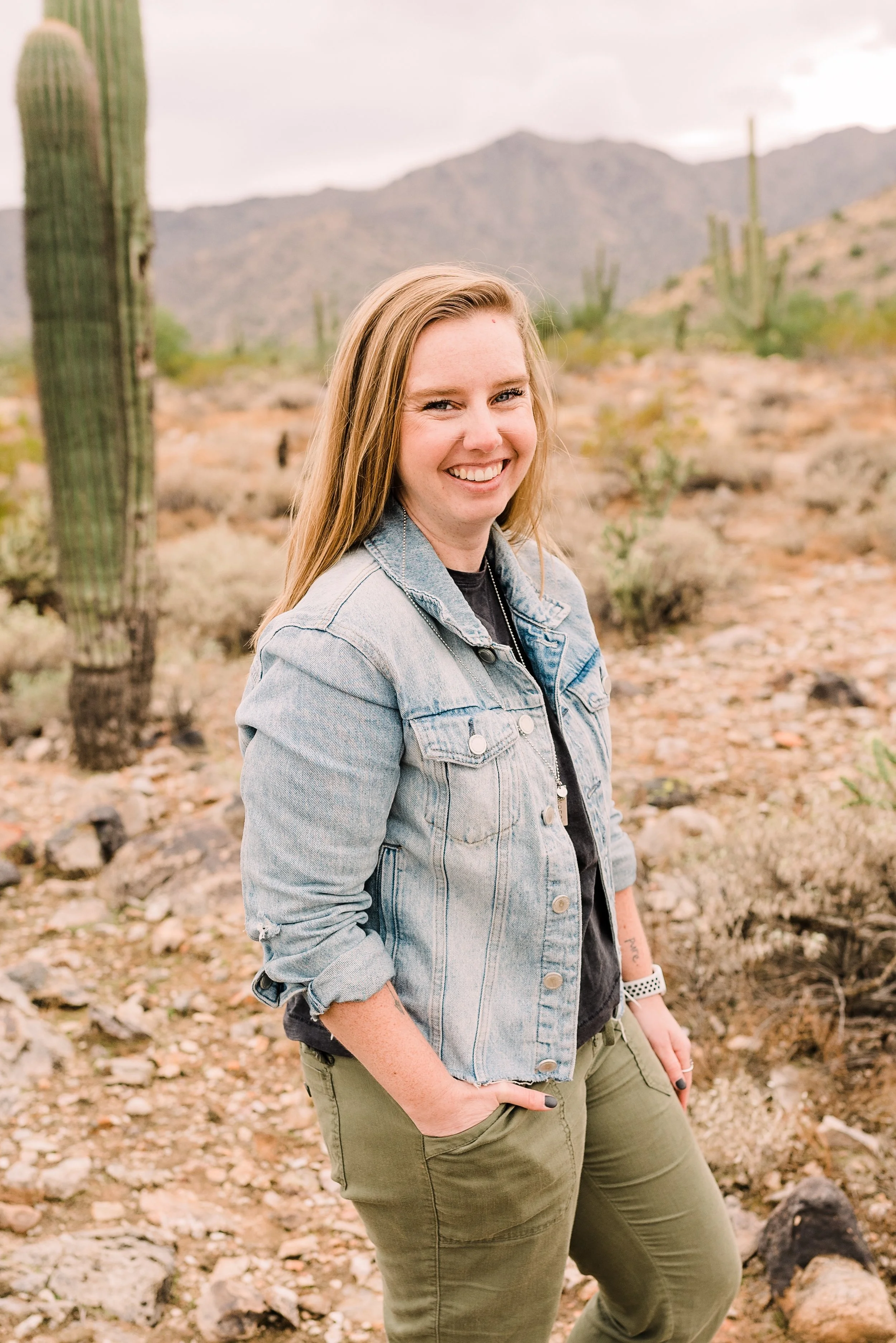 A young woman with long blond hair, wearing a light blue denim jacket and olive green pants, smiling with one hand in her pocket in a desert landscape with mountains, cacti, and rocky ground.