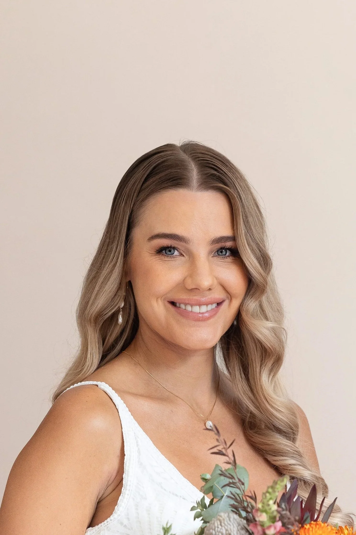 A smiling woman with long, wavy blonde hair smiling, wearing a white sleeveless dress, pearl earrings, a delicate necklace.