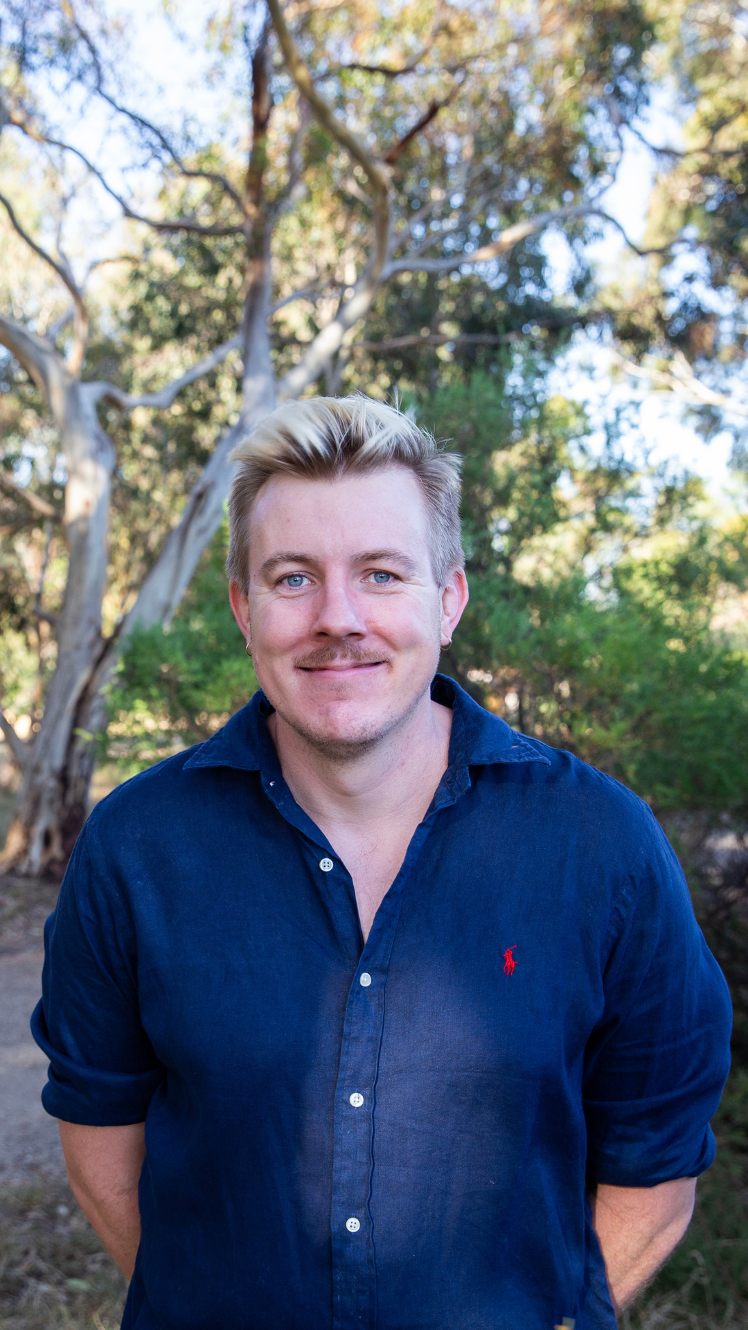 A man with blonde hair, blue eyes, and a light mustache standing outdoors with trees in the background, smiling at the camera, wearing a dark blue button-up shirt.