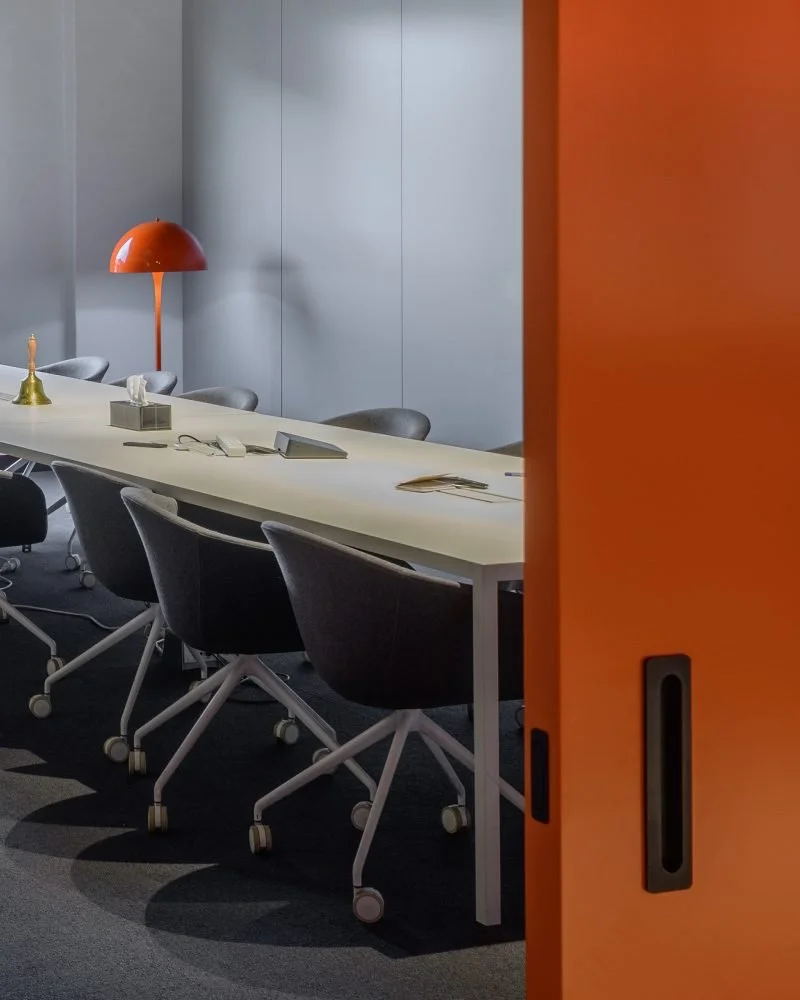 An empty conference room with a long white table, black office chairs, a red-orange lamp, a tissue box, and some papers, seen through an orange sliding door.