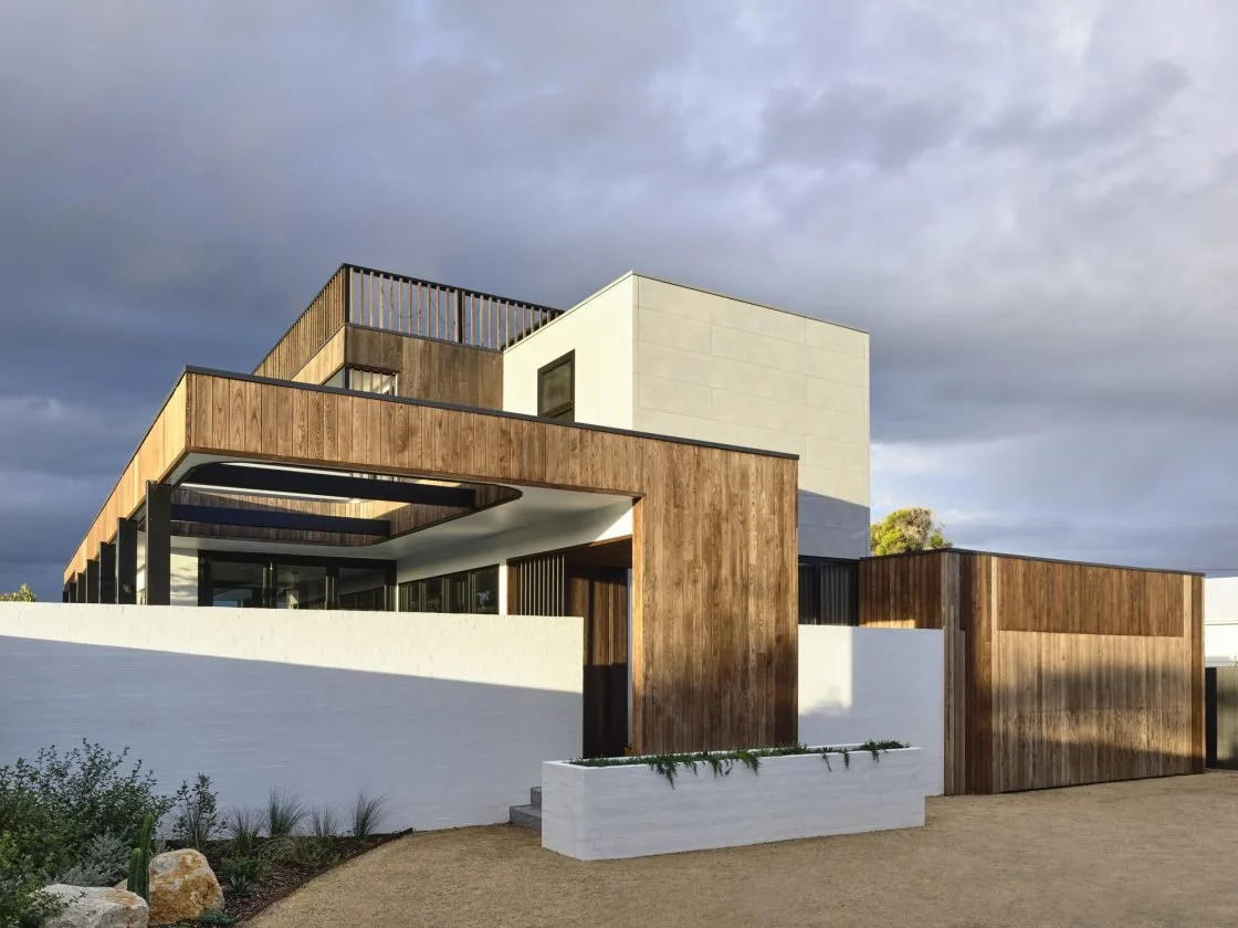 Modern house with a mix of wood, white concrete, and black accents, under a cloudy sky, featuring clean geometric architecture and outdoor landscaping.