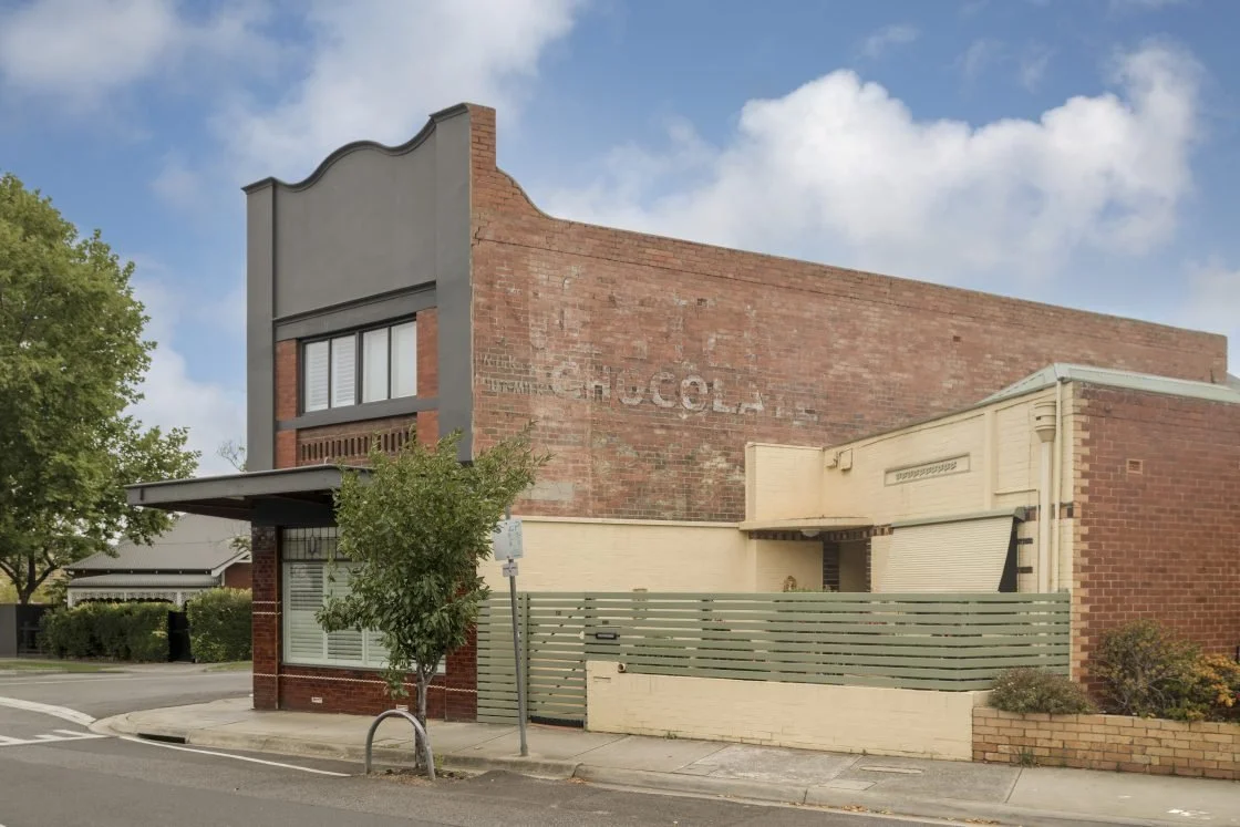A building with a brick facade and a faded sign that reads 'Coca-Cola' on the side, situated on a street corner with trees and sidewalk.