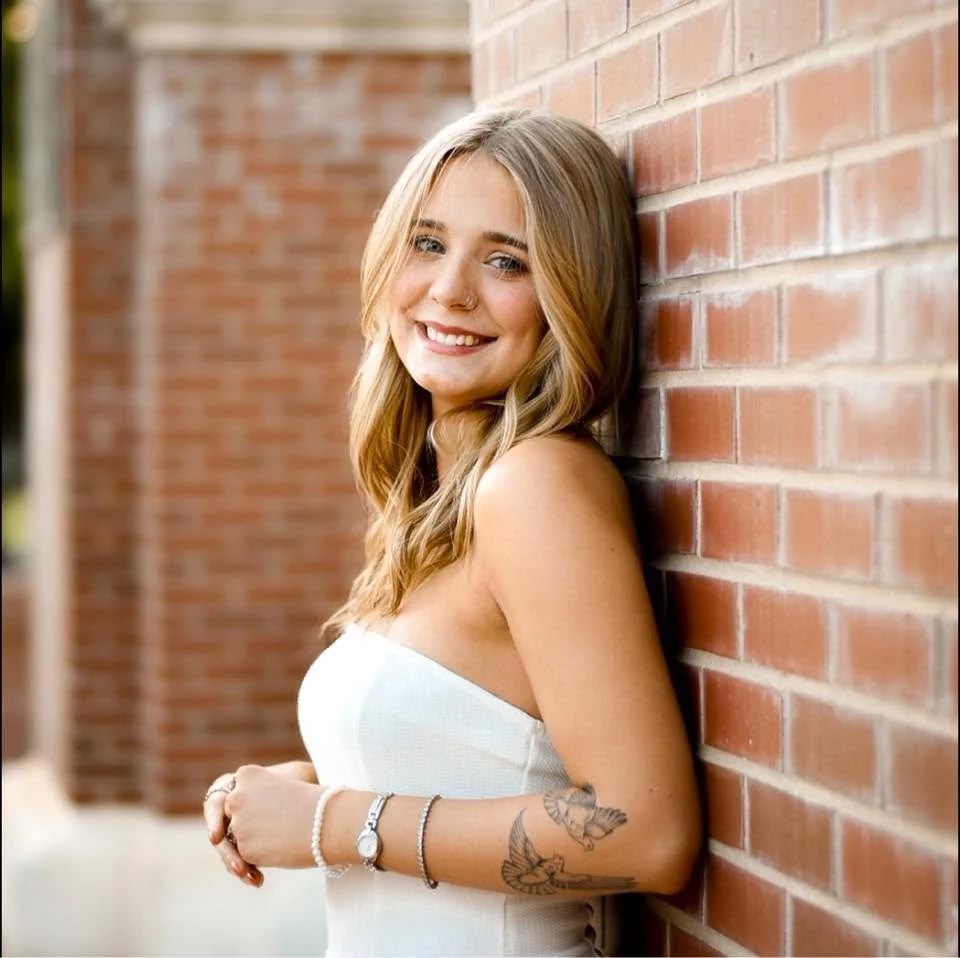 Smiling young woman with long wavy blonde hair wearing a white strapless top, standing against a brick wall outdoors.