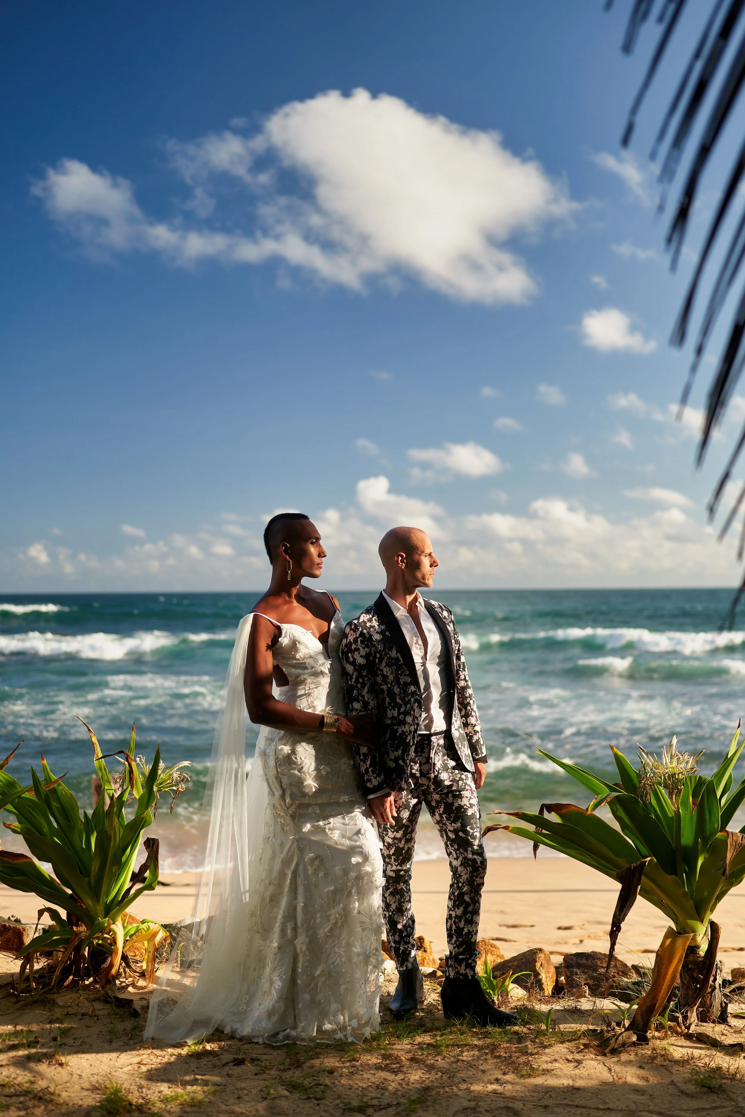 A queer couple in wedding attire stands on a beach, with the ocean and a partly cloudy sky in the background. The bride wears a white wedding gown, and the groom wears a floral suit, with greenery nearby.