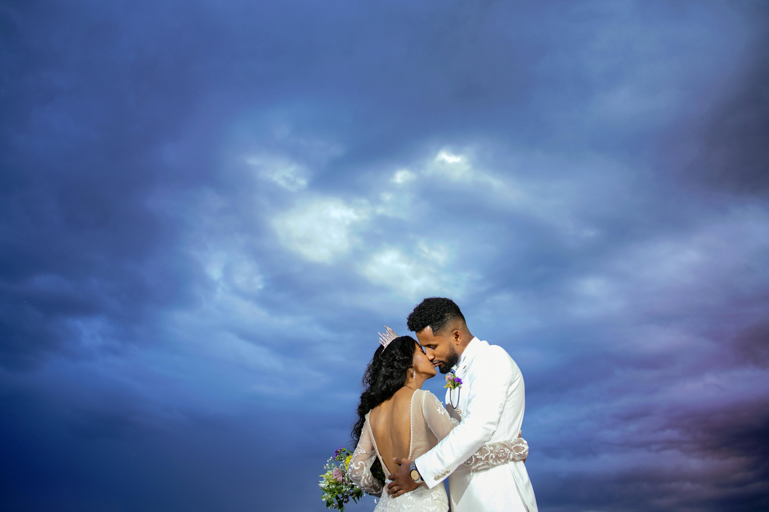 A bride and groom embracing and touching foreheads against a cloudy sky sky at sunset.
