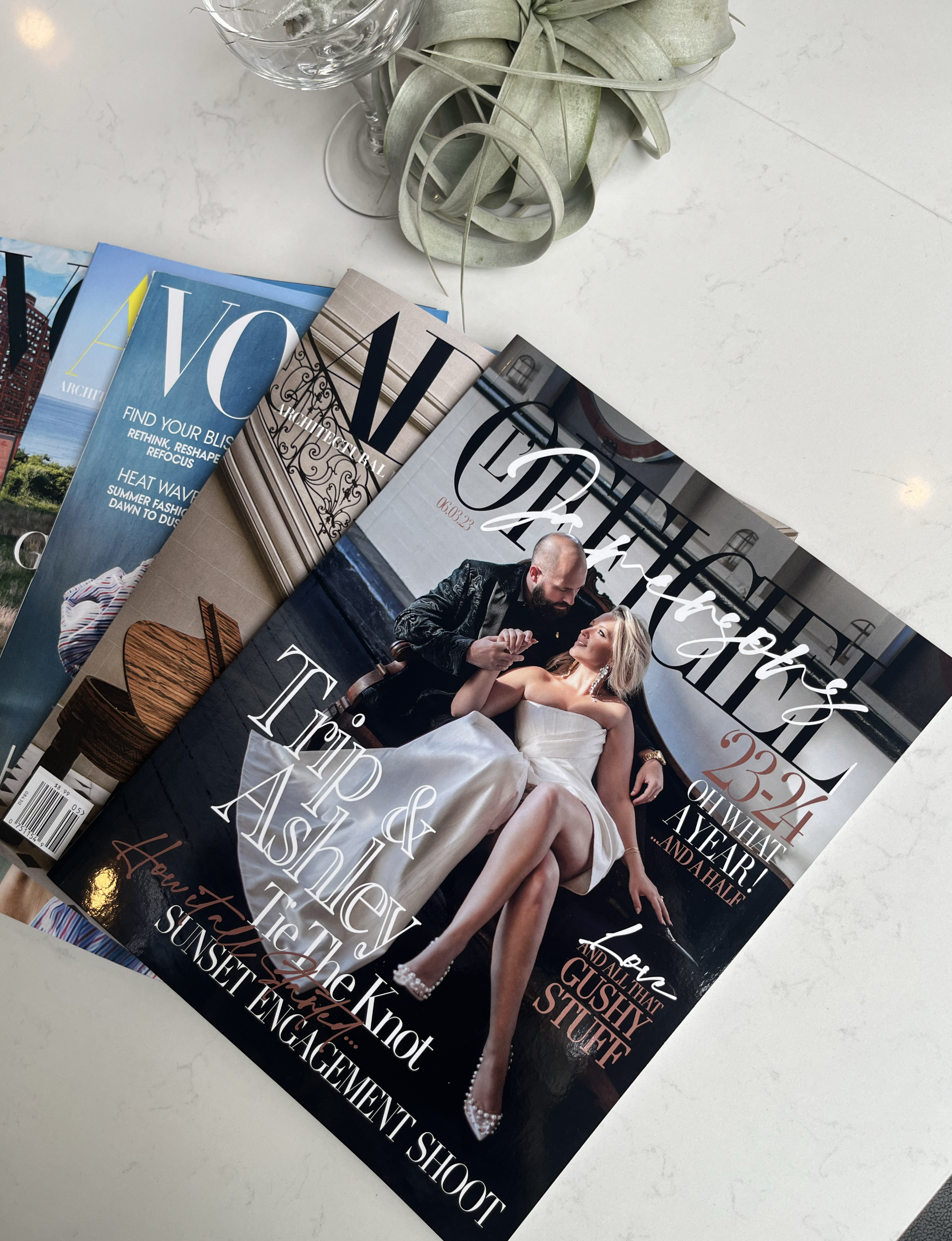 A wedding magazine titled 'The Enchanted' with a cover photo of a bride in a white dress and a groom in a black suit sitting on a bench, with other magazine issues partially visible underneath on a white marble surface. There is a glass of water and a plant with green leaves in a round pot nearby.
