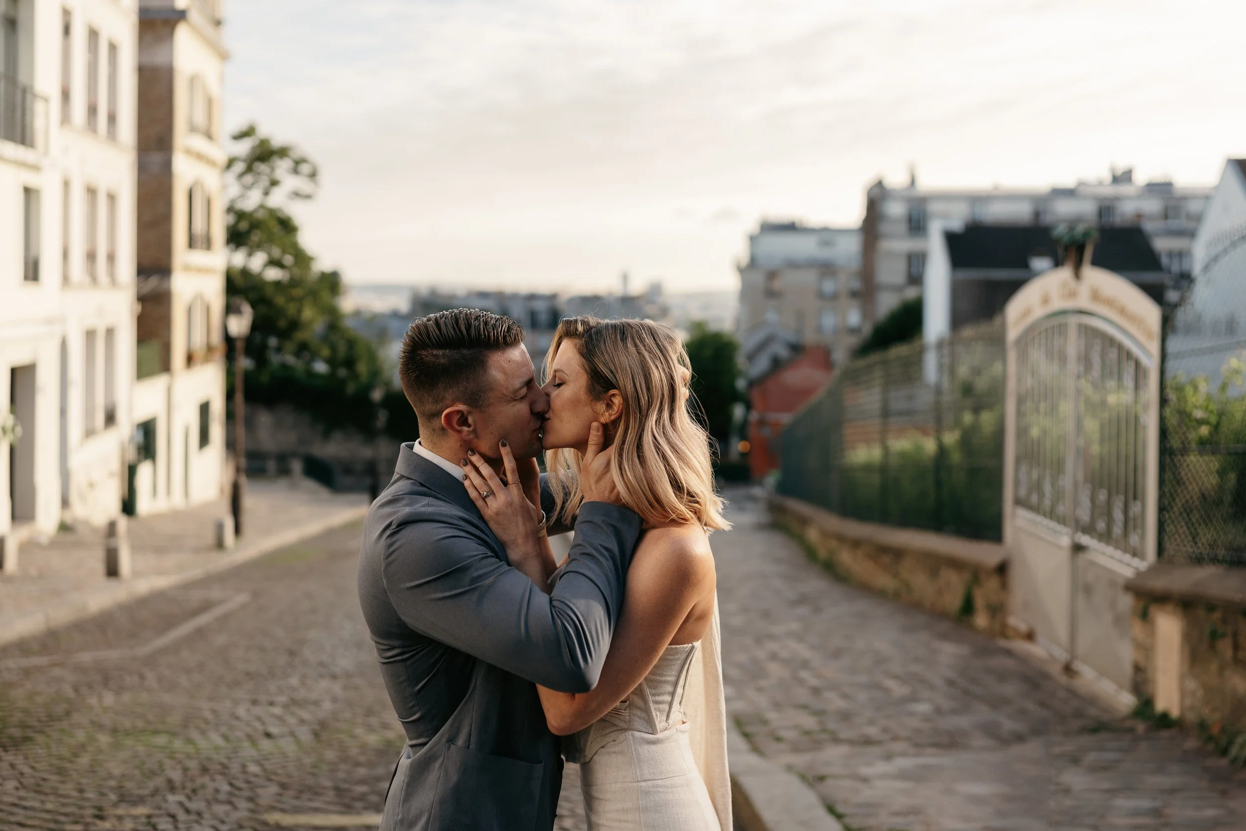 A couple shares a kiss on a cobblestone street in Paris, with buildings and a fence in the background during sunset.