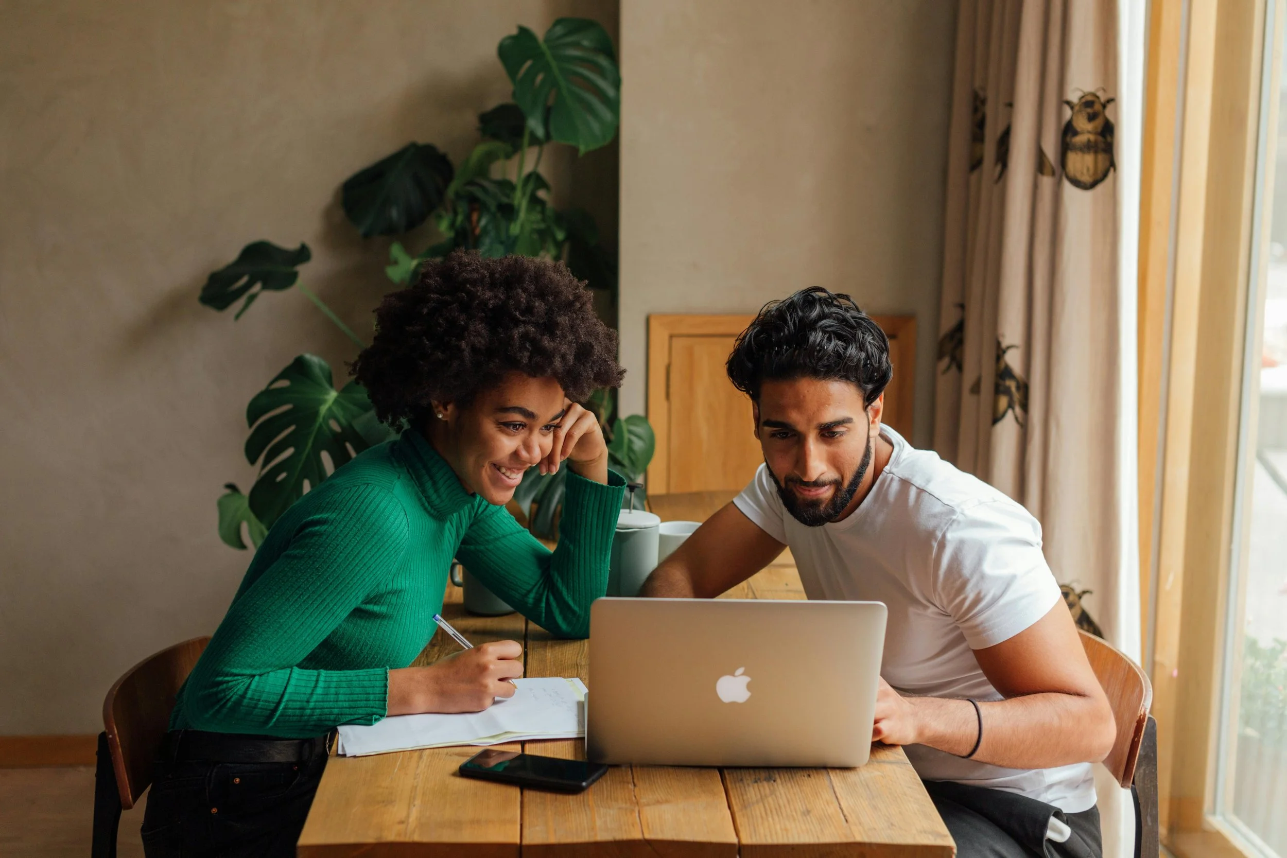 A woman and a man are sitting at a wooden table, looking at a laptop and smiling. The woman has curly hair and is wearing a green sweater, while the man has dark hair and is wearing a white t-shirt. They are in a room with a large window, beige curtains with owl designs, and a large green plant in the background.