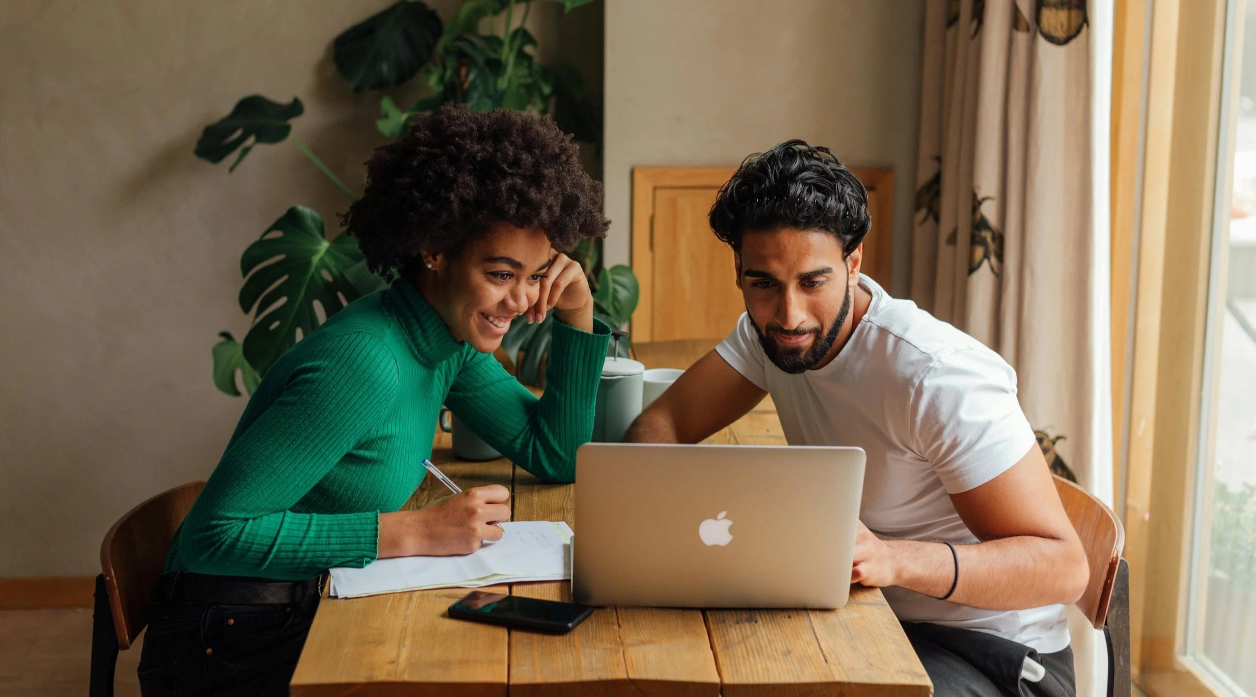A man and woman sitting at a wooden table looking at a laptop screen, with notebooks and a smartphone on the table.
