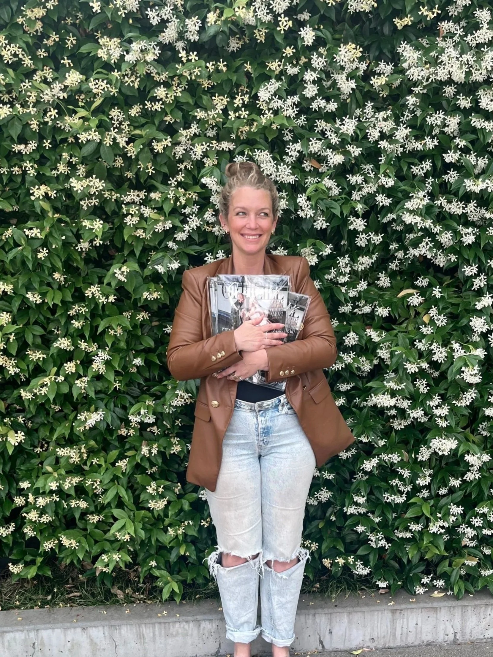 A woman with light skin, blonde hair in a bun, smiling, stands in front of a wall of green leaves with white flowers, holding several magazines or newspapers close to her chest.