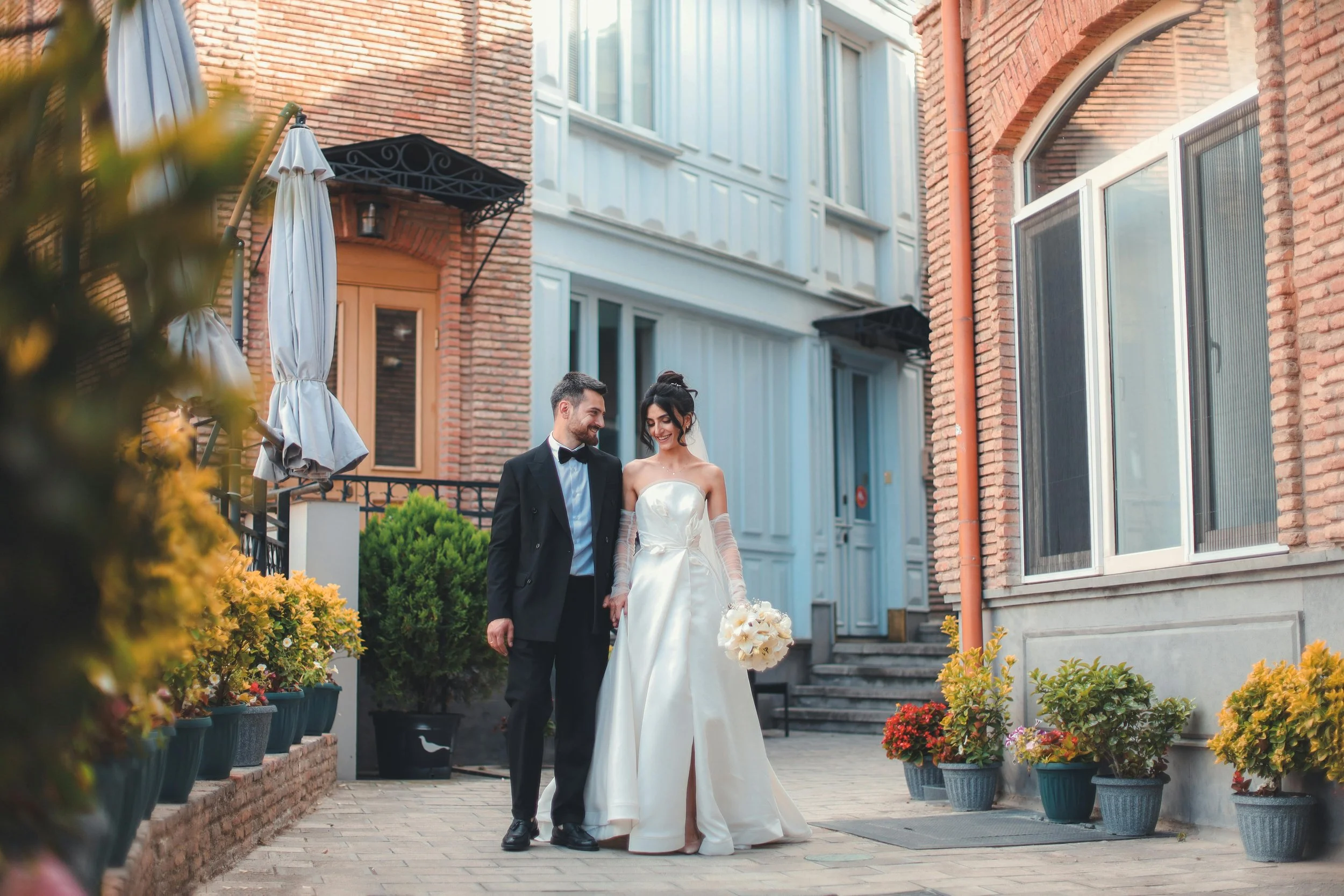 A bride and groom walking hand in hand outside of a brick building, smiling, on their wedding day. The bride wears a strapless white wedding gown and the groom is dressed in a black tuxedo with a bow tie. She holds a bouquet of white flowers.