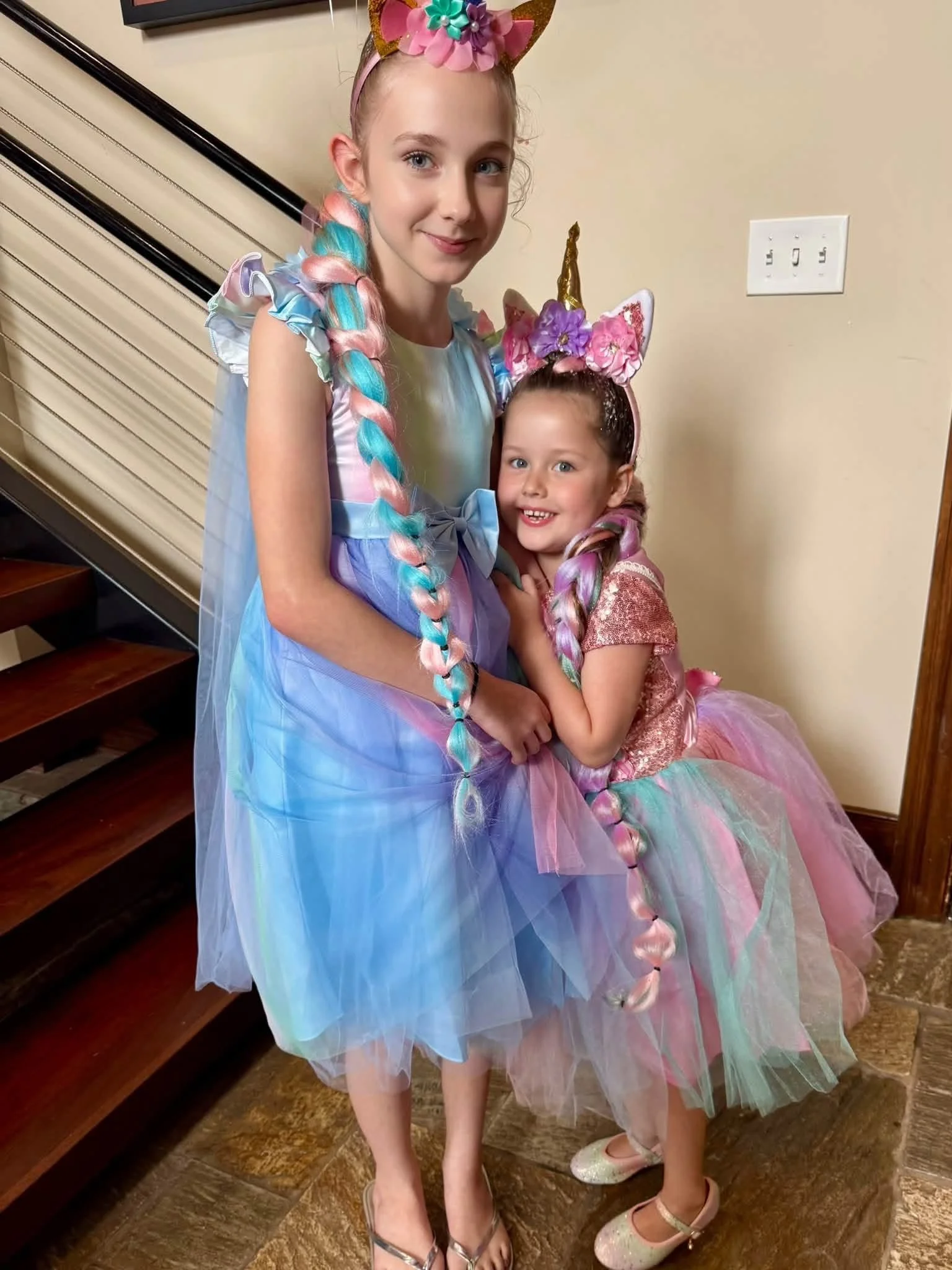 Two young girls dressed as unicorns in colorful tutus with unicorn headbands, standing close together indoors.