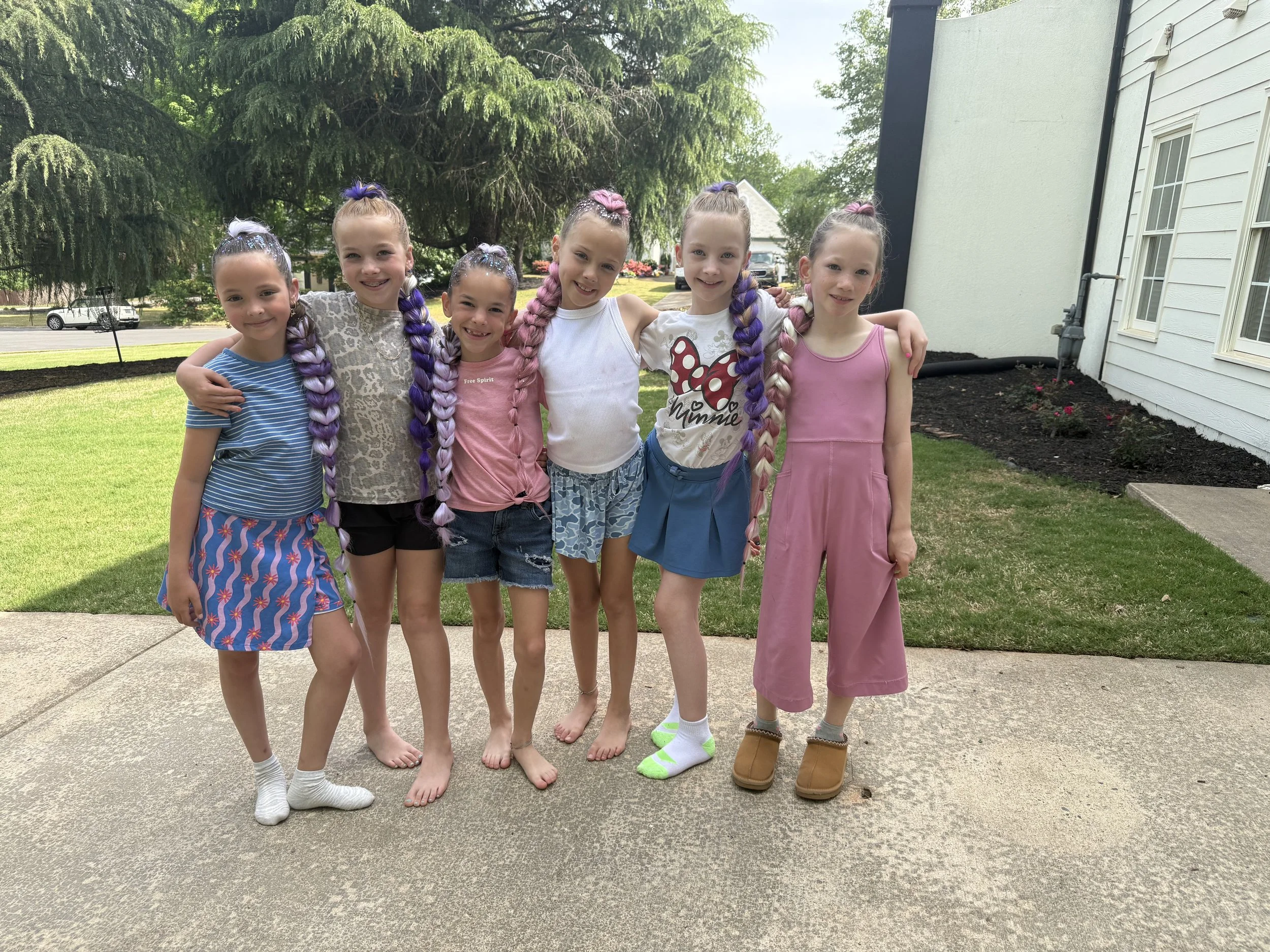 A group of six young girls standing outside on a driveway with trees and a house in the background. They are smiling and have colorful braided hair, wearing casual summer clothes and some barefoot.