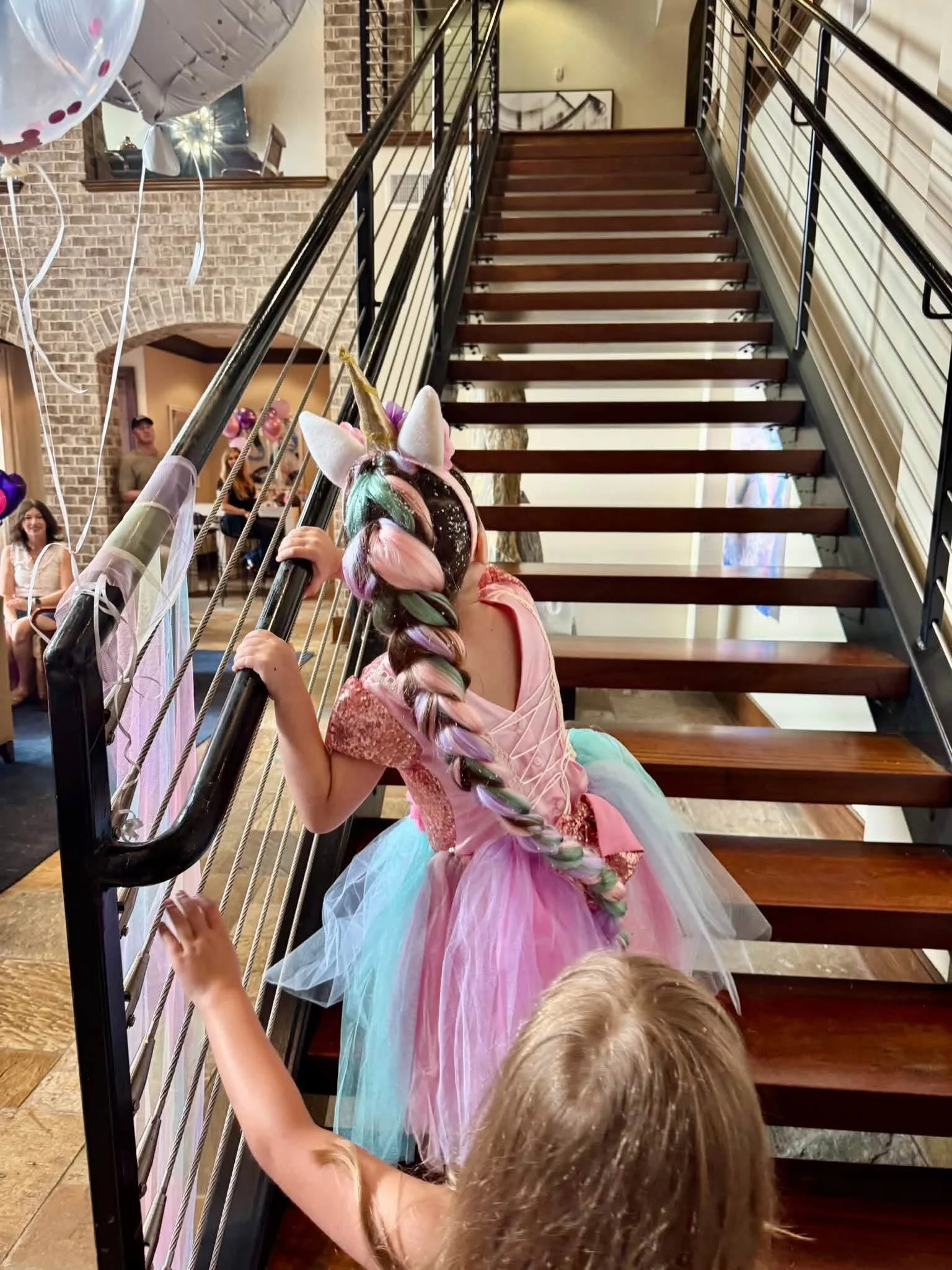 Child dressed as a unicorn princess at a birthday party, wearing a pink dress and a colorful unicorn horn headband, holding onto a staircase railing, with balloons in the background.