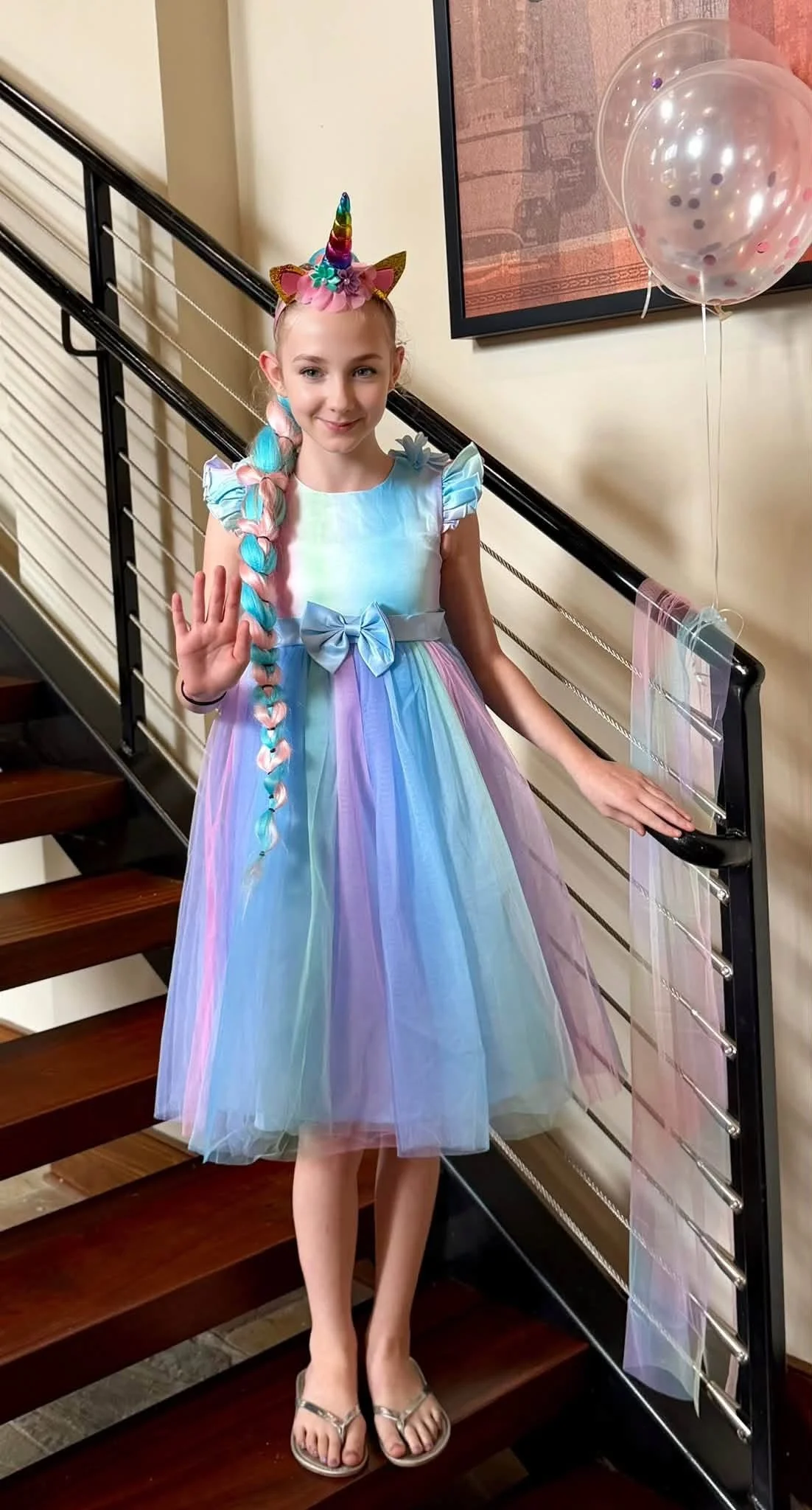 A young girl wearing a pastel rainbow-colored tulle dress with ruffled sleeves and a bow on the waist, standing on a staircase. She has long, multicolored braided hair, a unicorn headband, and is waving with her left hand. There are pink balloons with confetti inside floating beside her.