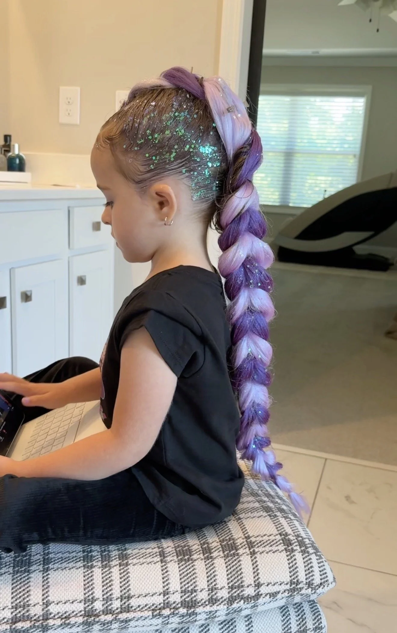 A young girl with long purple and pink braid hairstyle with glitter, sitting on a plaid cushion and using a laptop in a bright room with a window and a white cabinet in the background.