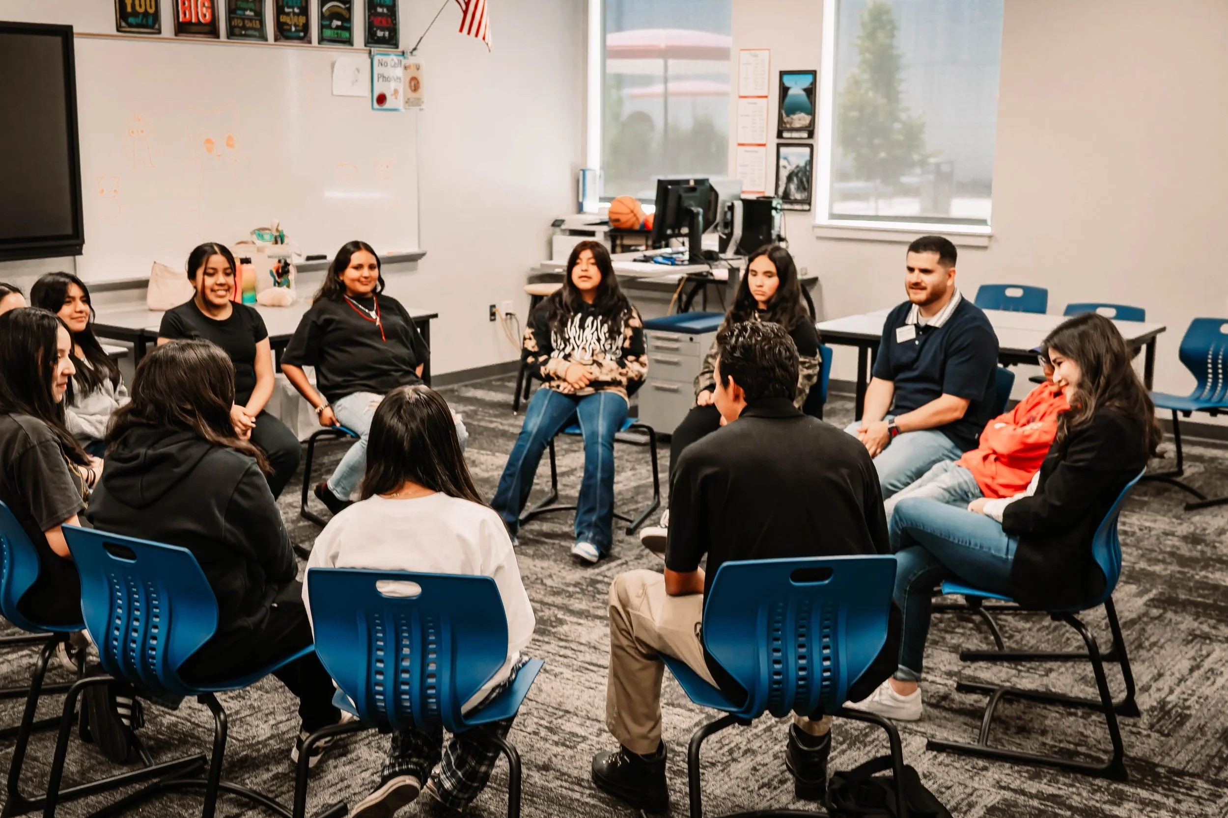 A group of mostly youth and a couple adults sit in a circle of chairs in a classroom.