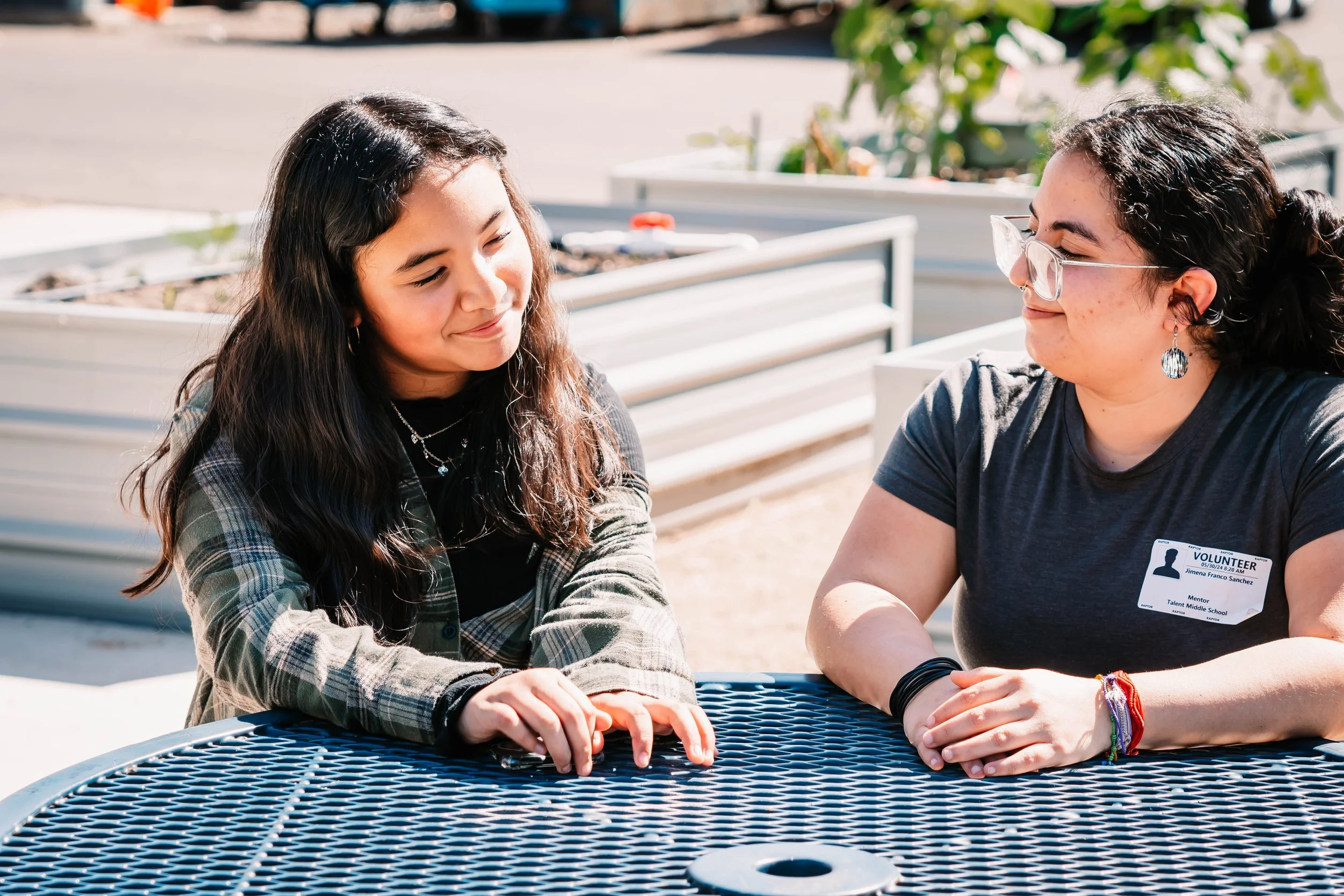 A Latina adult and young person sit at a table together outdoors, planter boxes in the background.