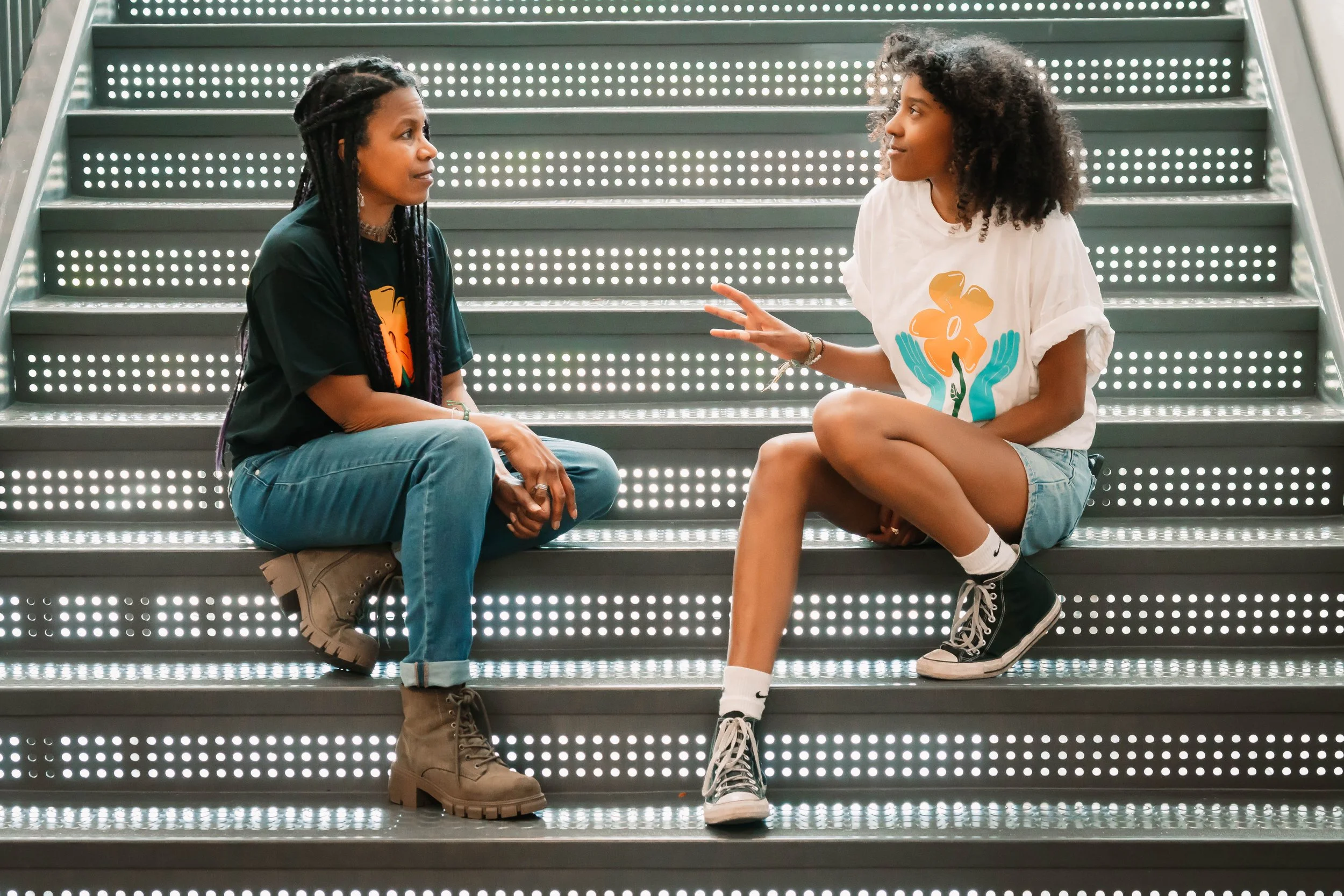 A Black adult and Black youth sit on a flight of stairs. The adult listens while the young person speaks.