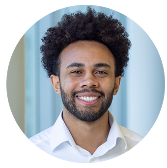 Close-up of a smiling young man with curly hair and a beard, wearing a white shirt, standing indoors with a blurred background.
