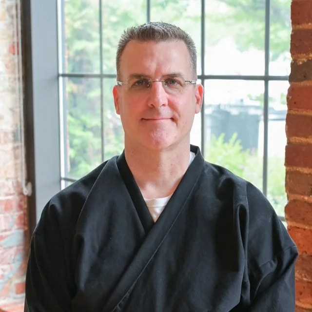 A man with glasses and short hair wearing a black robe standing indoors near a large window with a brick wall on the side.
