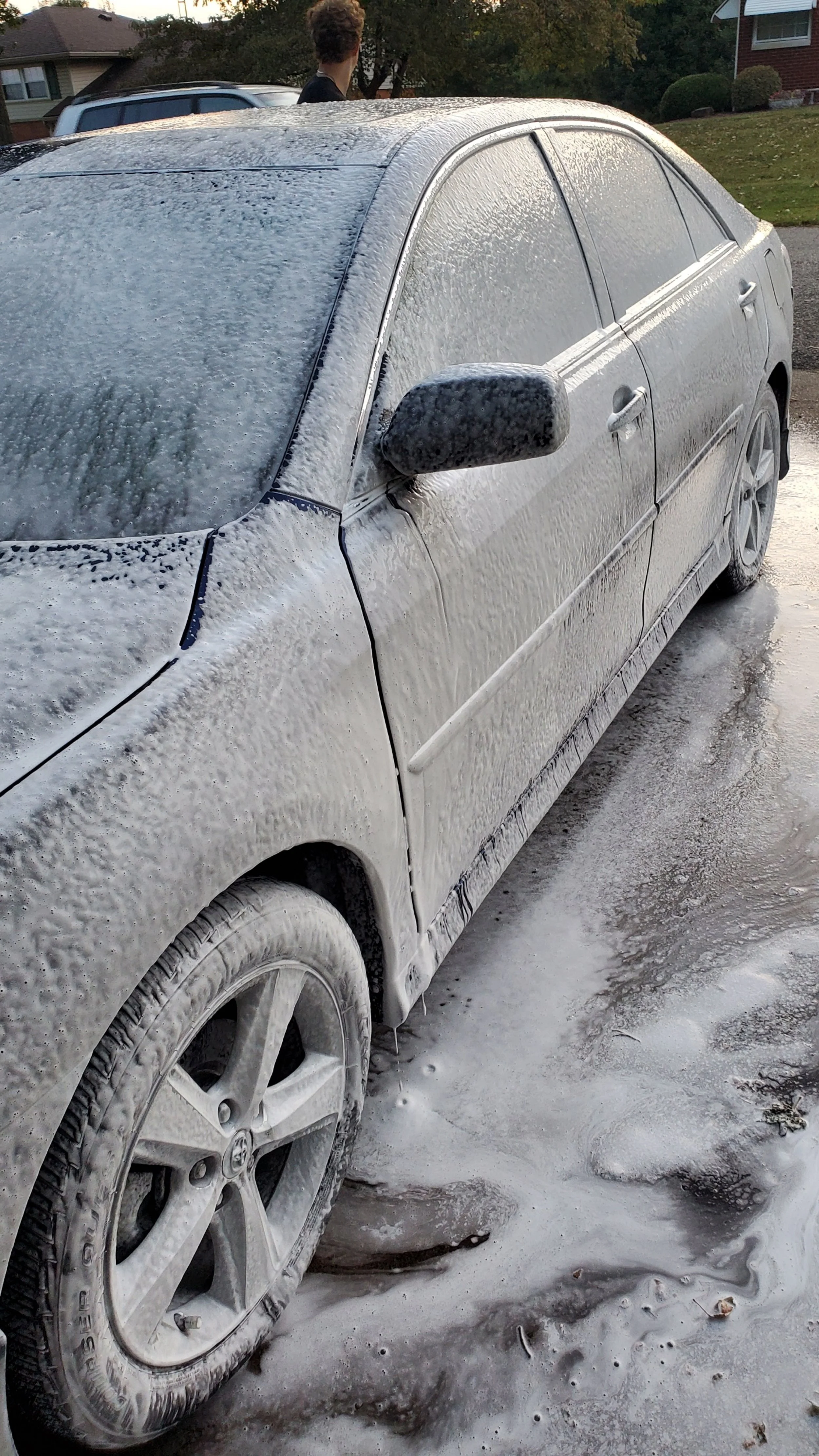 A silver sedan covered in soap and foam during car washing, with a person in the background.